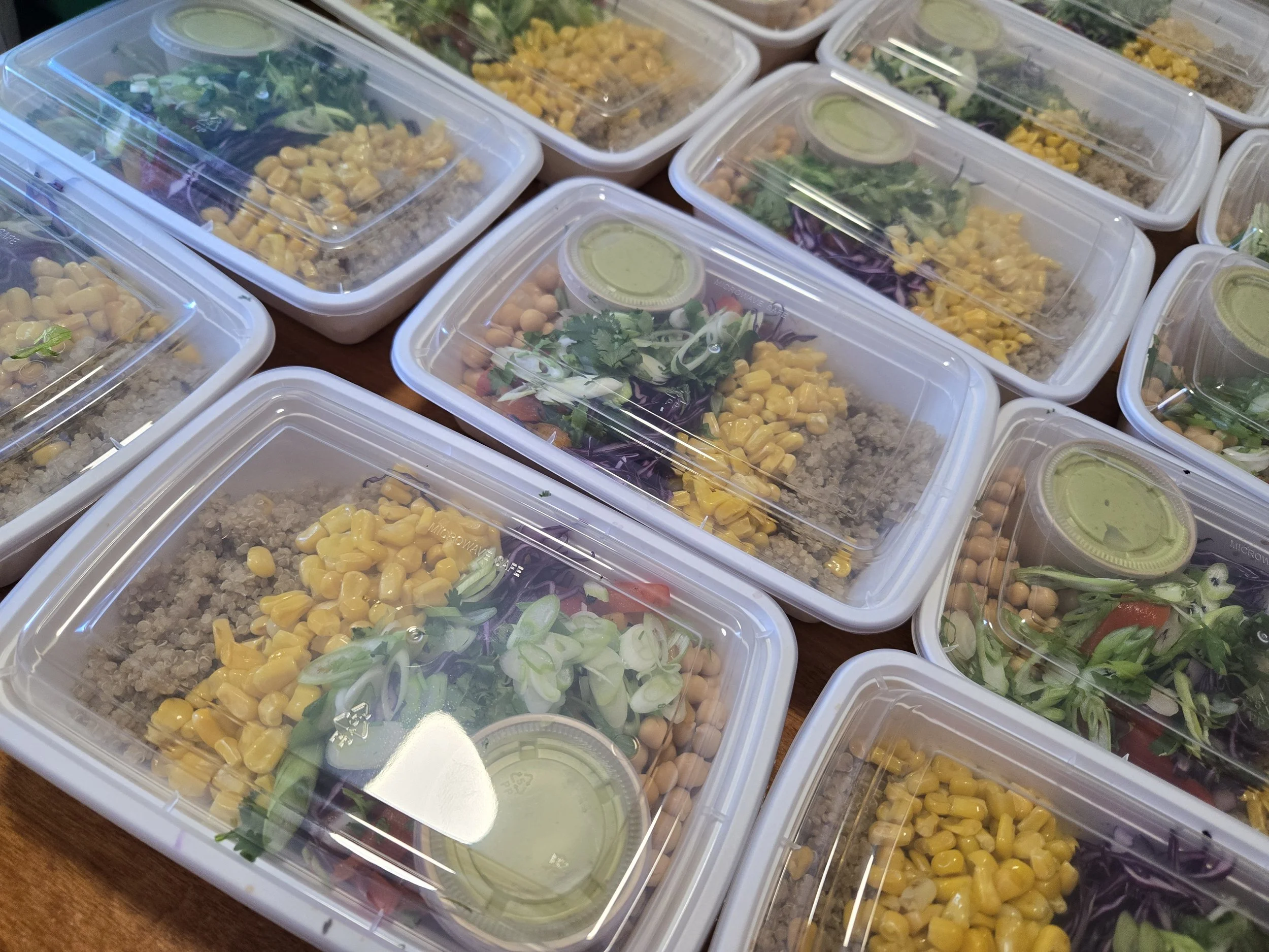 Multiple plastic containers filled with salads and grain dishes, topped with transparent plastic lids, placed on a wooden surface.