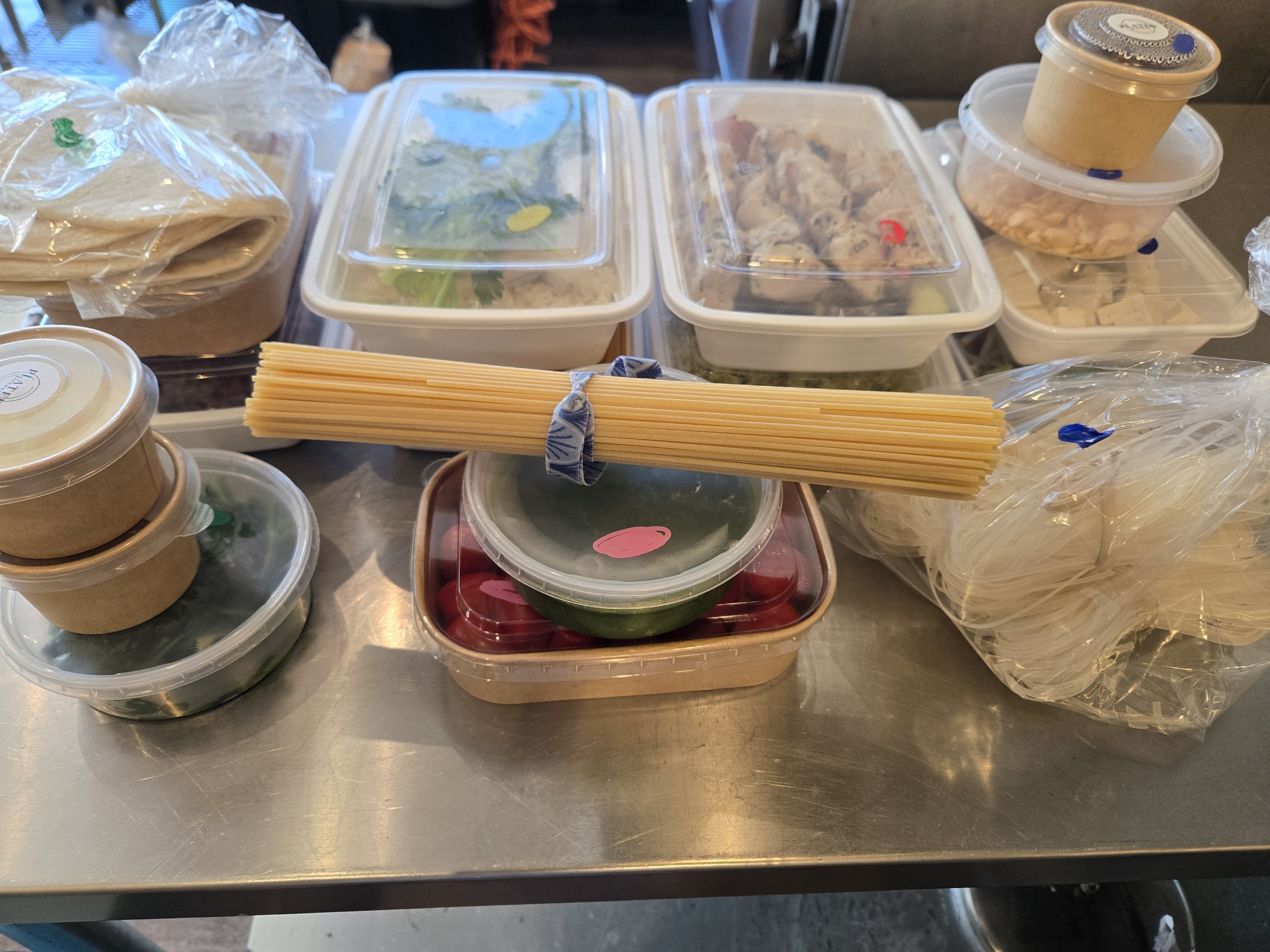 Various containers of prepped ingredients and uncooked pasta on a stainless steel surface.