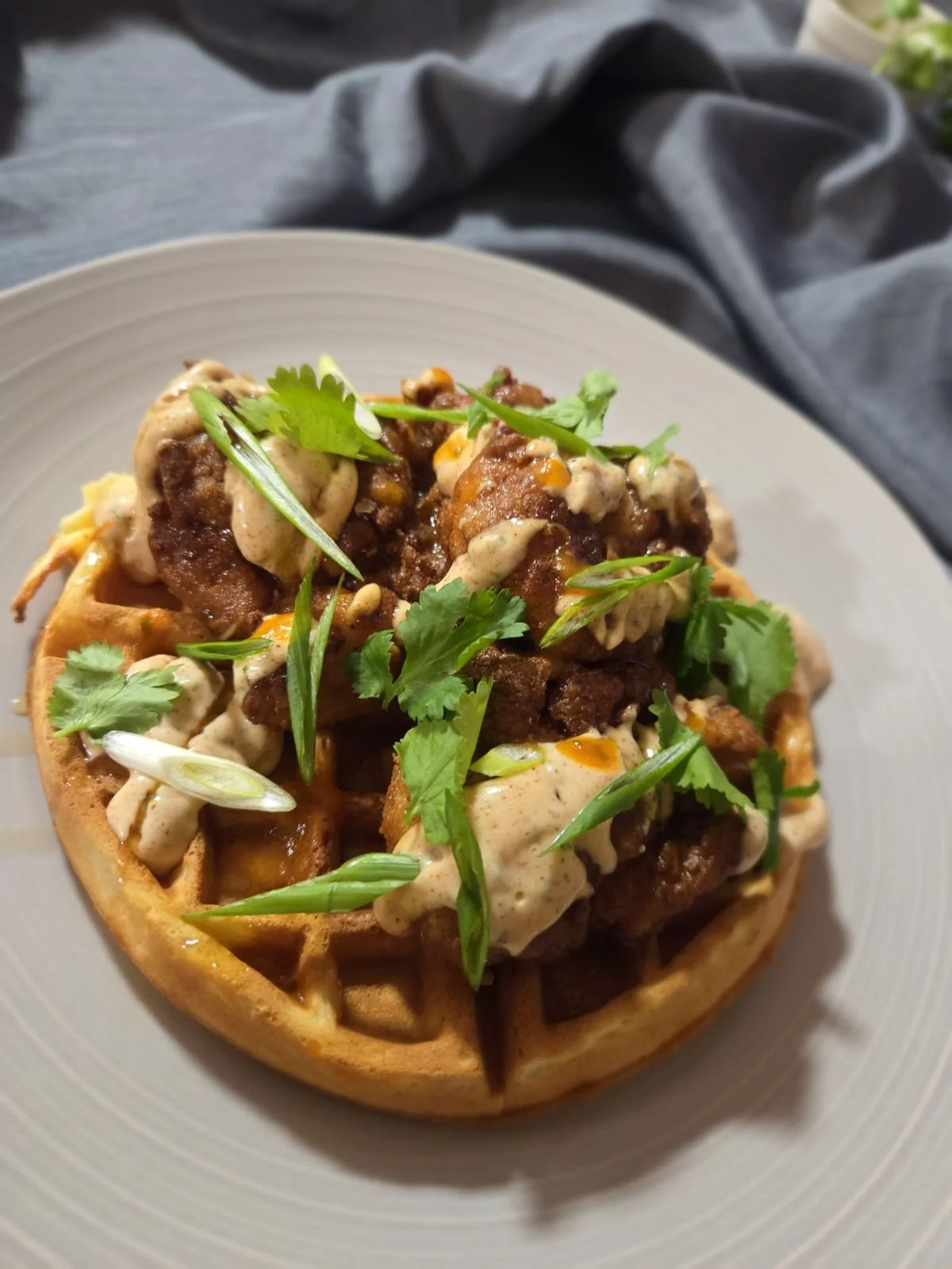 A waffle topped with fried chicken, scallions, cilantro, maple syrup, and a creamy sauce.