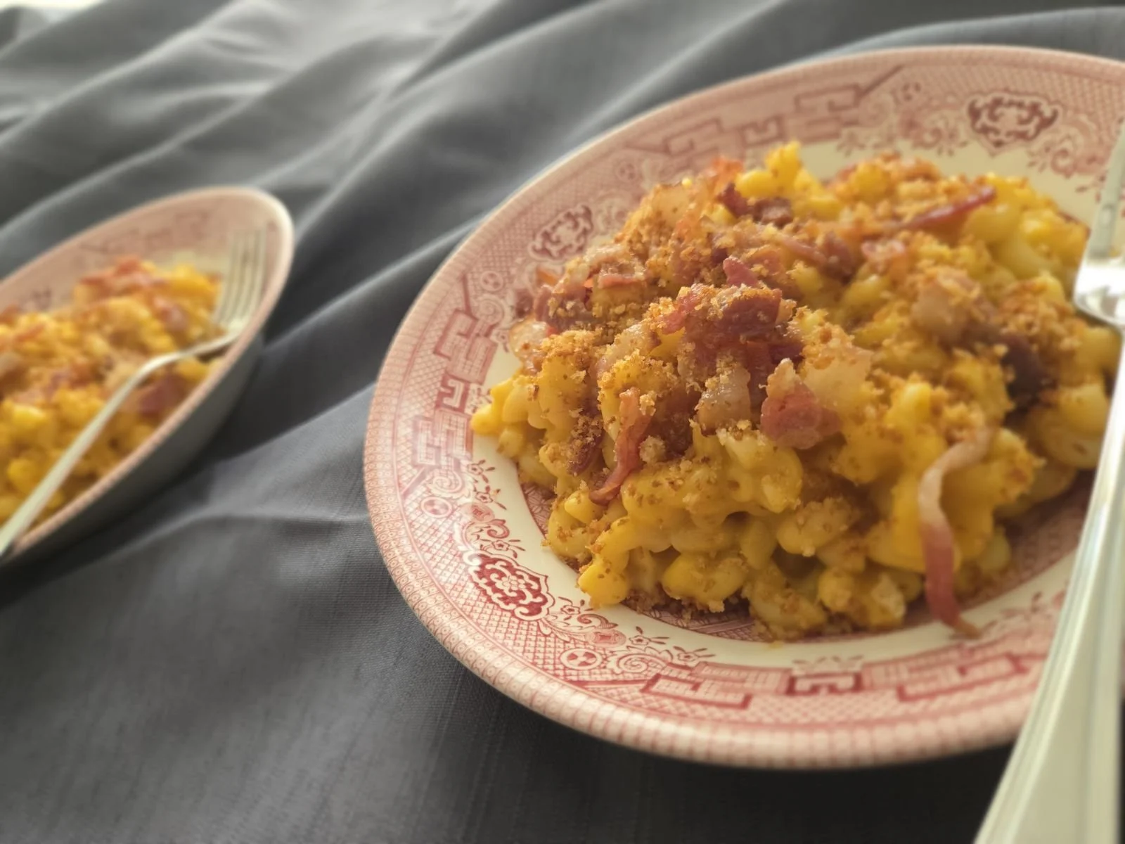 Close-up of a serving of macaroni and cheese with bacon bits on a decorative pink and white plate, with two similar dishes in the background.