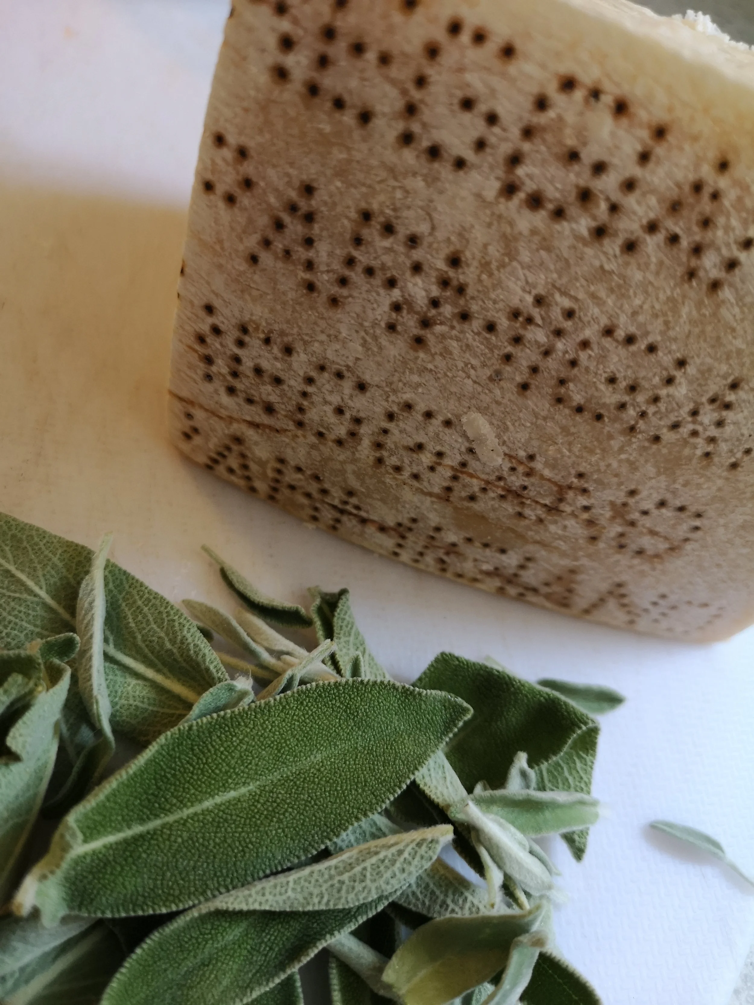 Close-up of a round, beige, textured cheese with small holes, and a sprig of green sage leaves nearby.