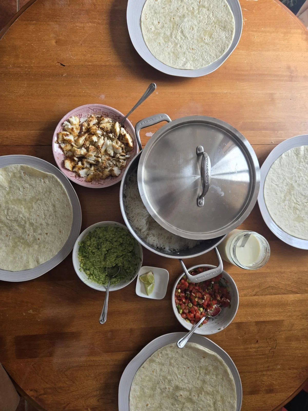A traditional Mexican meal setup with tortillas, shredded chicken, chopped vegetables, guacamole, lime wedges, rice, salsa, and a mason jar of creme fraiche on a wooden table.