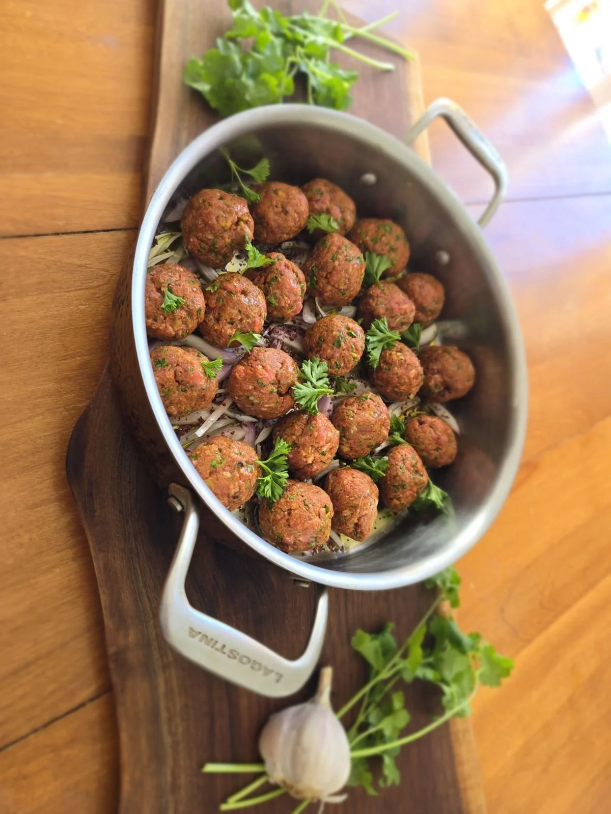 Meatballs garnished with parsley in a metal container on a wooden tray, with garlic and parsley on a wooden table.
