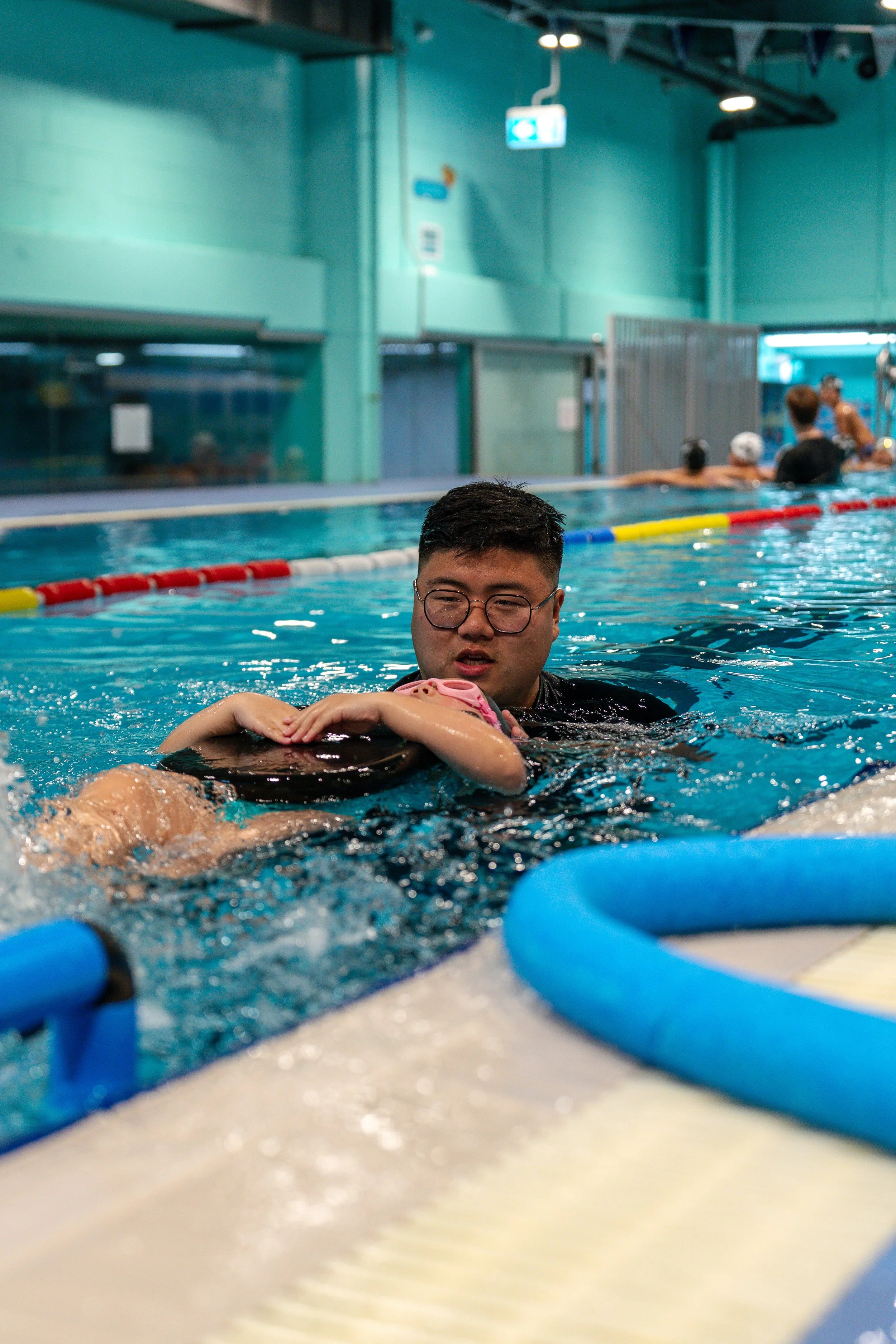 Young man wearing glasses and black swimsuit holding a young girl in a swimming pool, with other swimmers and a teal-colored indoor pool facility in the background.