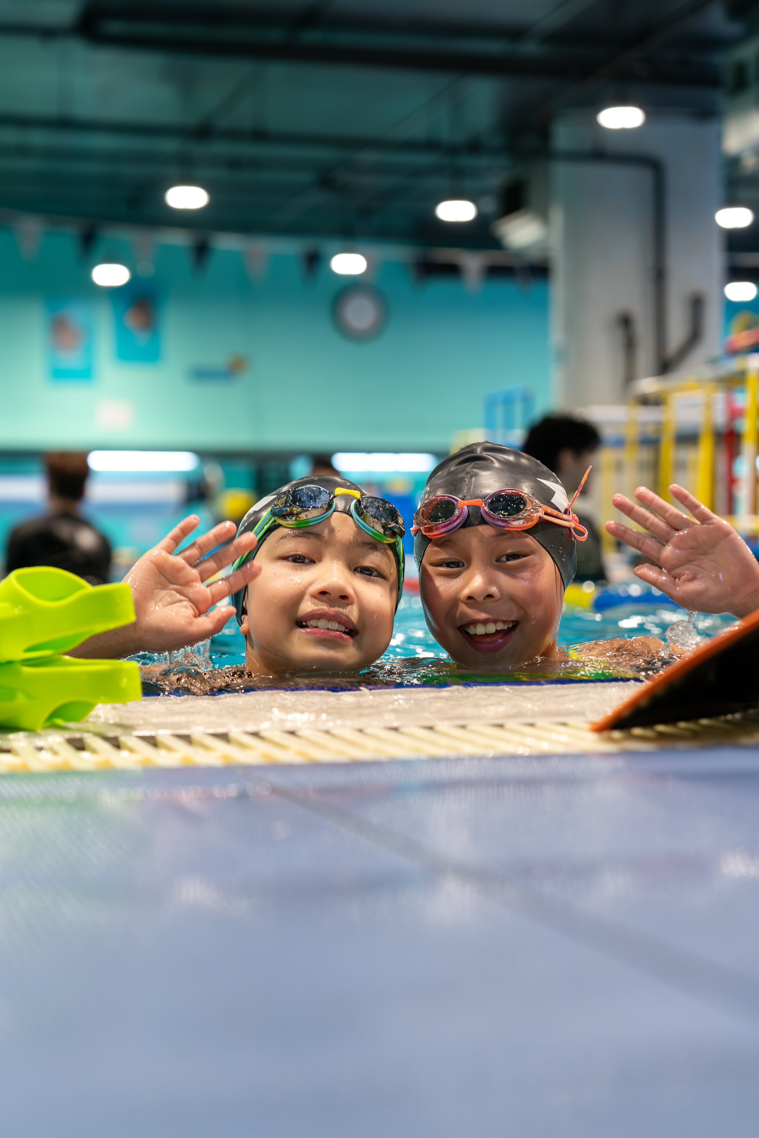 Two children in swimming goggles and swim caps smiling and waving in a swimming pool.