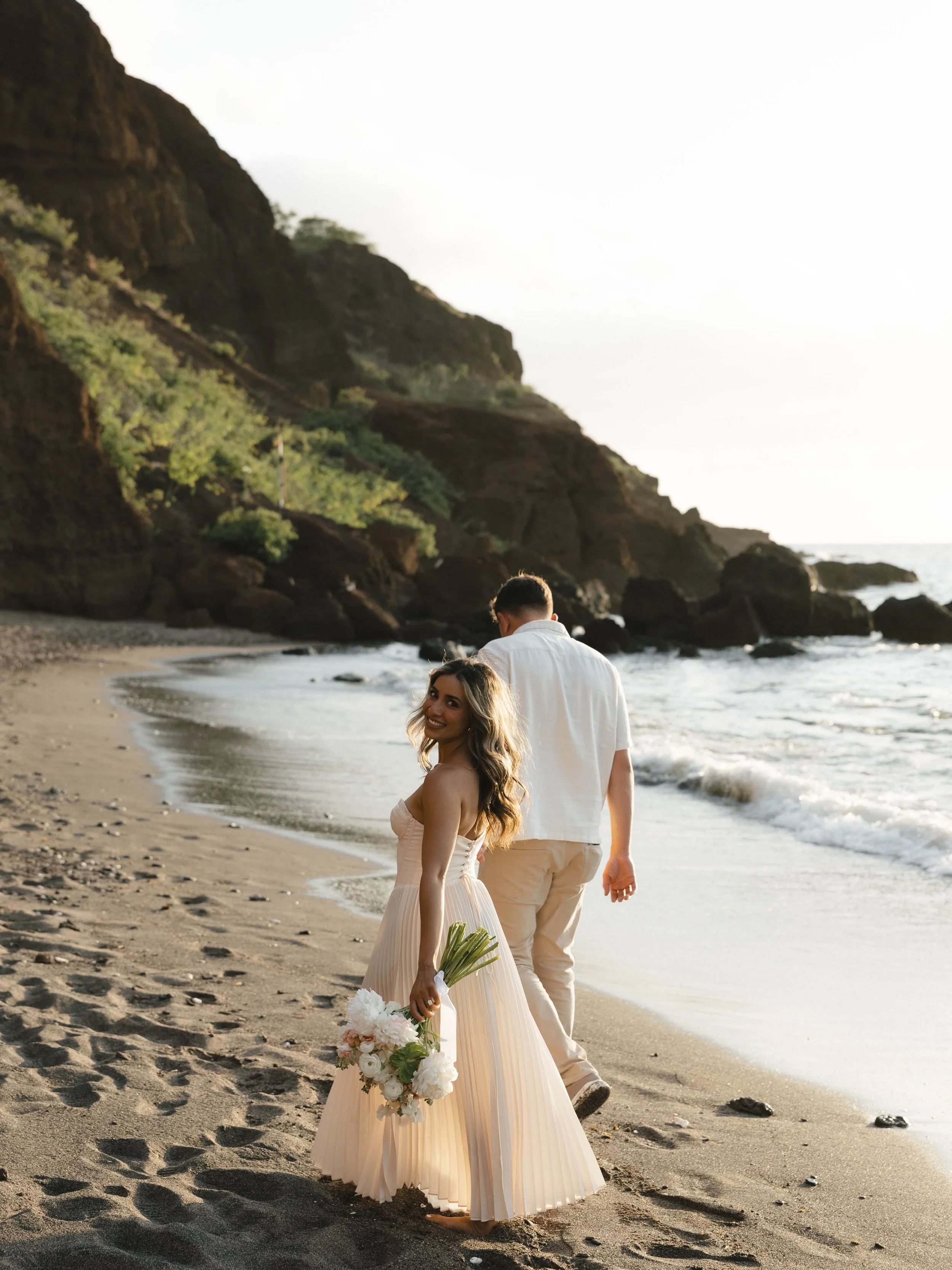 A woman in a light pink, strapless, pleated dress holding a bouquet of white flowers stands barefoot on the beach, smiling at the camera. A man in a white shirt and beige pants walks ahead along the shoreline, back turned to the camera. The background features ocean waves, rocky shoreline, and steep cliffside with green vegetation.
