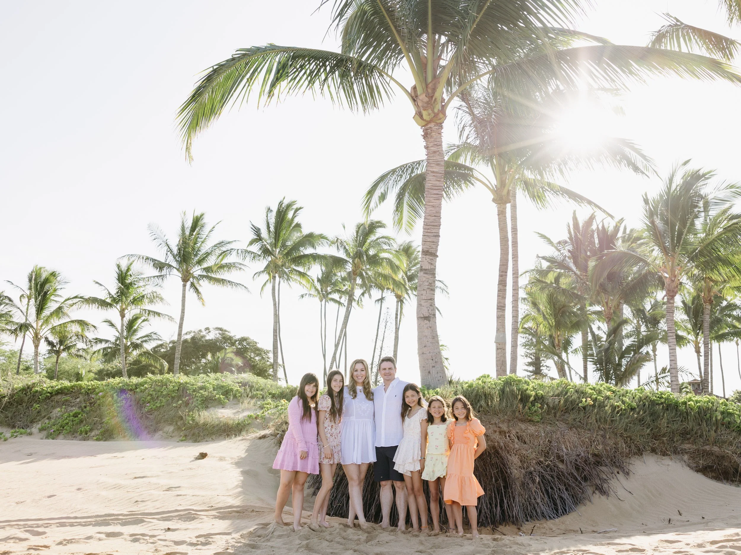 A family of seven people standing on a beach in front of a large palm tree, with additional palm trees and a sandy beach in the background. The sun is shining brightly, creating a lens flare effect.