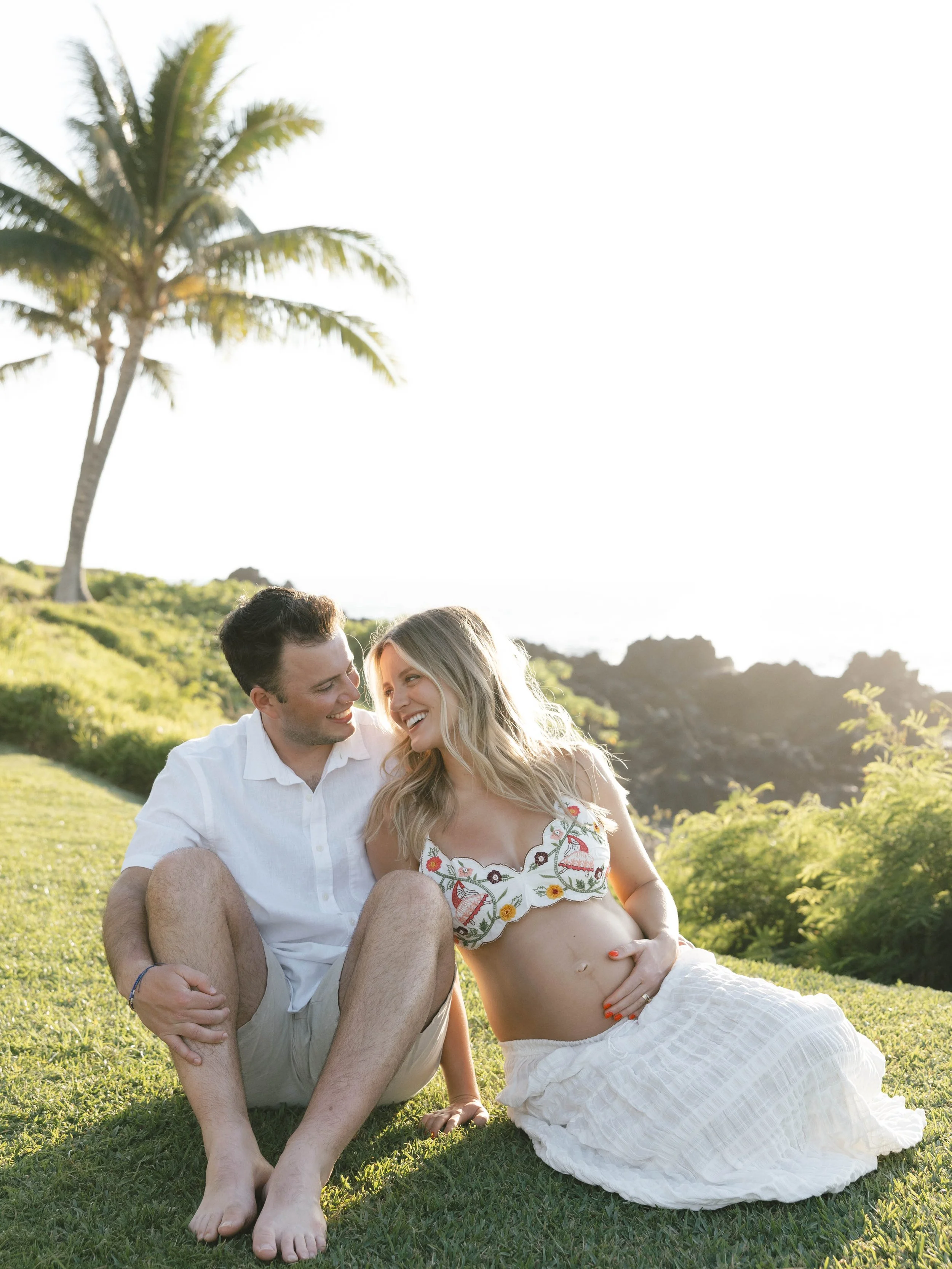 A couple sitting on grass outdoors, smiling and looking at each other, with a palm tree and mountains in the background under a bright sky.