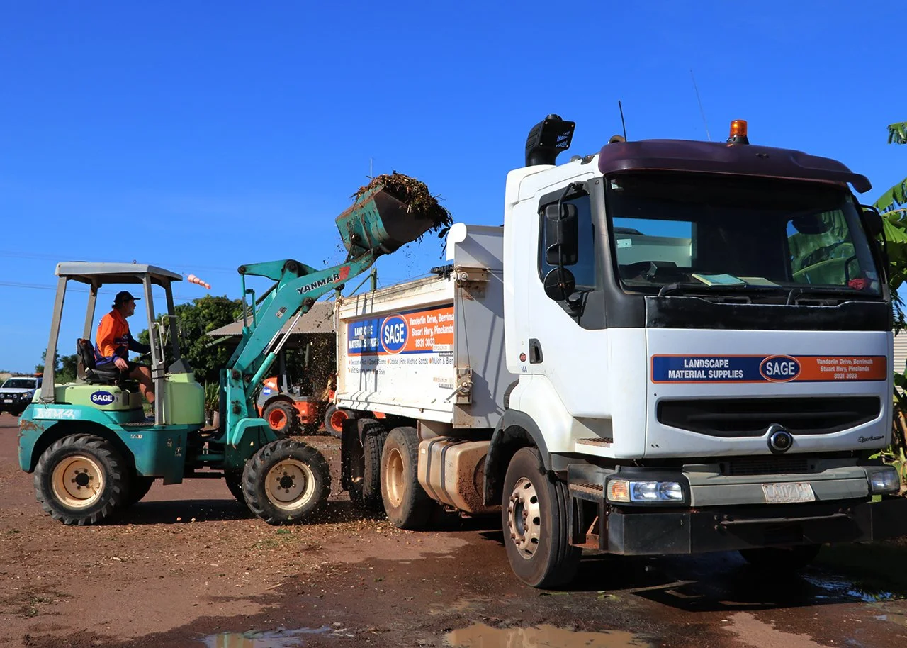 A worker operating a small green front loader is dumping soil or debris into the back of a white truck with a blue and orange logo. The truck has advertising for landscape material supplies.