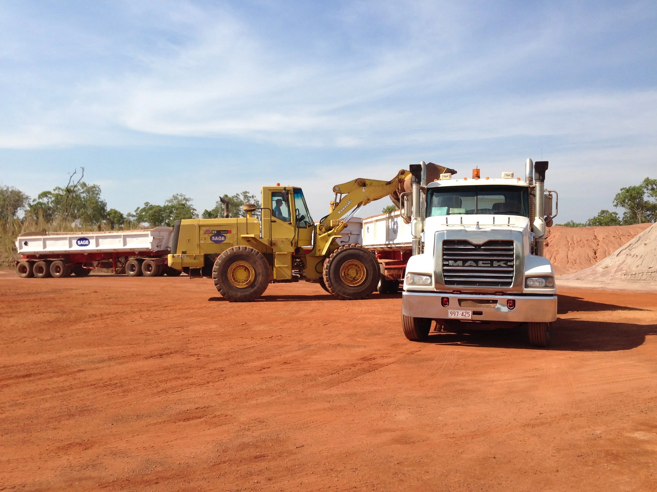 A construction site with a yellow excavator and a white Mack truck on reddish dirt, with trees and a partly cloudy sky in the background.