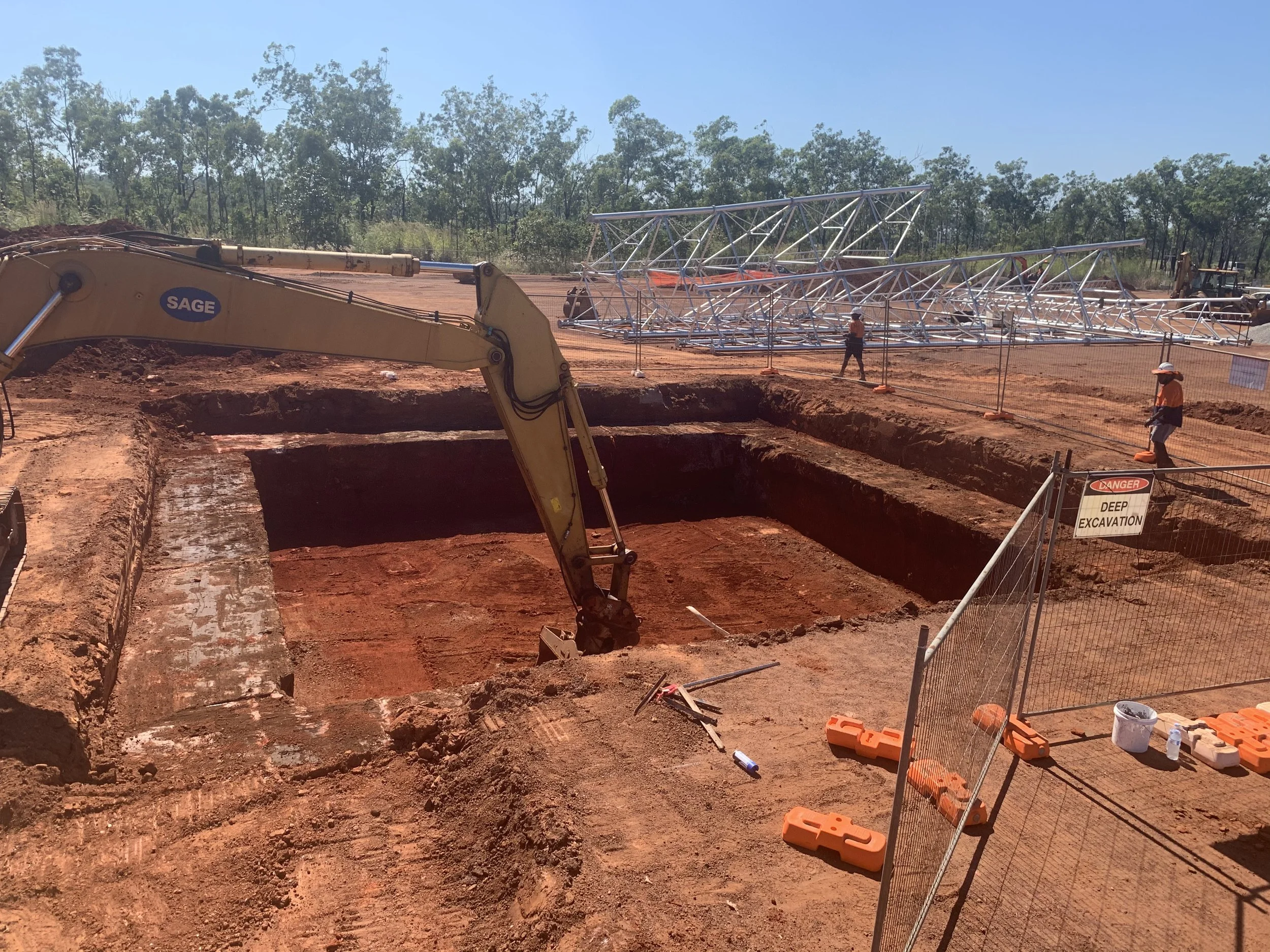 Construction site with deep excavation trench, metal framework, construction workers, and heavy machinery under a clear sky.