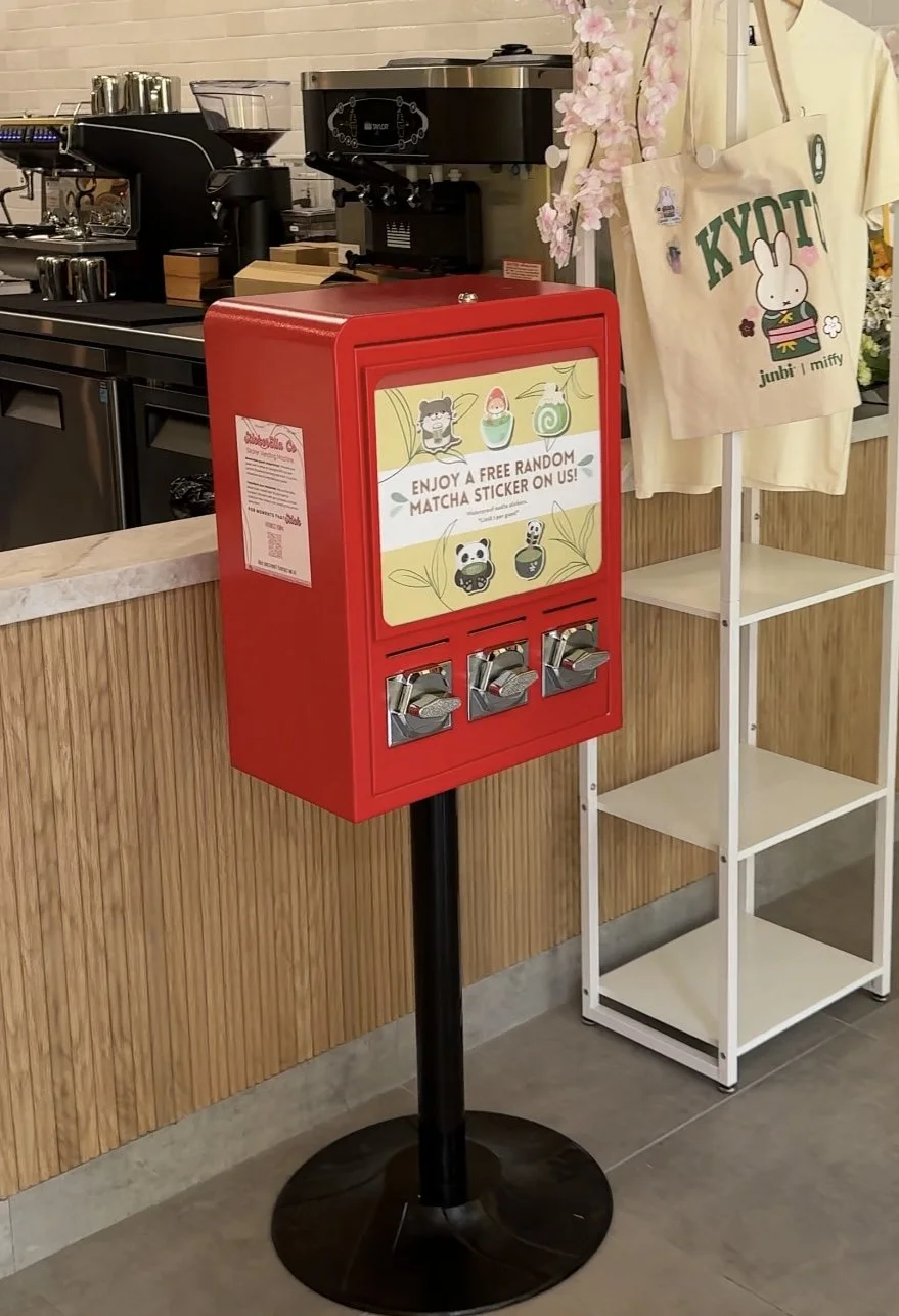 A red donation box with a sign offering free random matcha stickers, located indoors near a café counter with coffee equipment and a shelving unit with a tote bag that has a bunny illustration and the word 'KYOTI' on it.