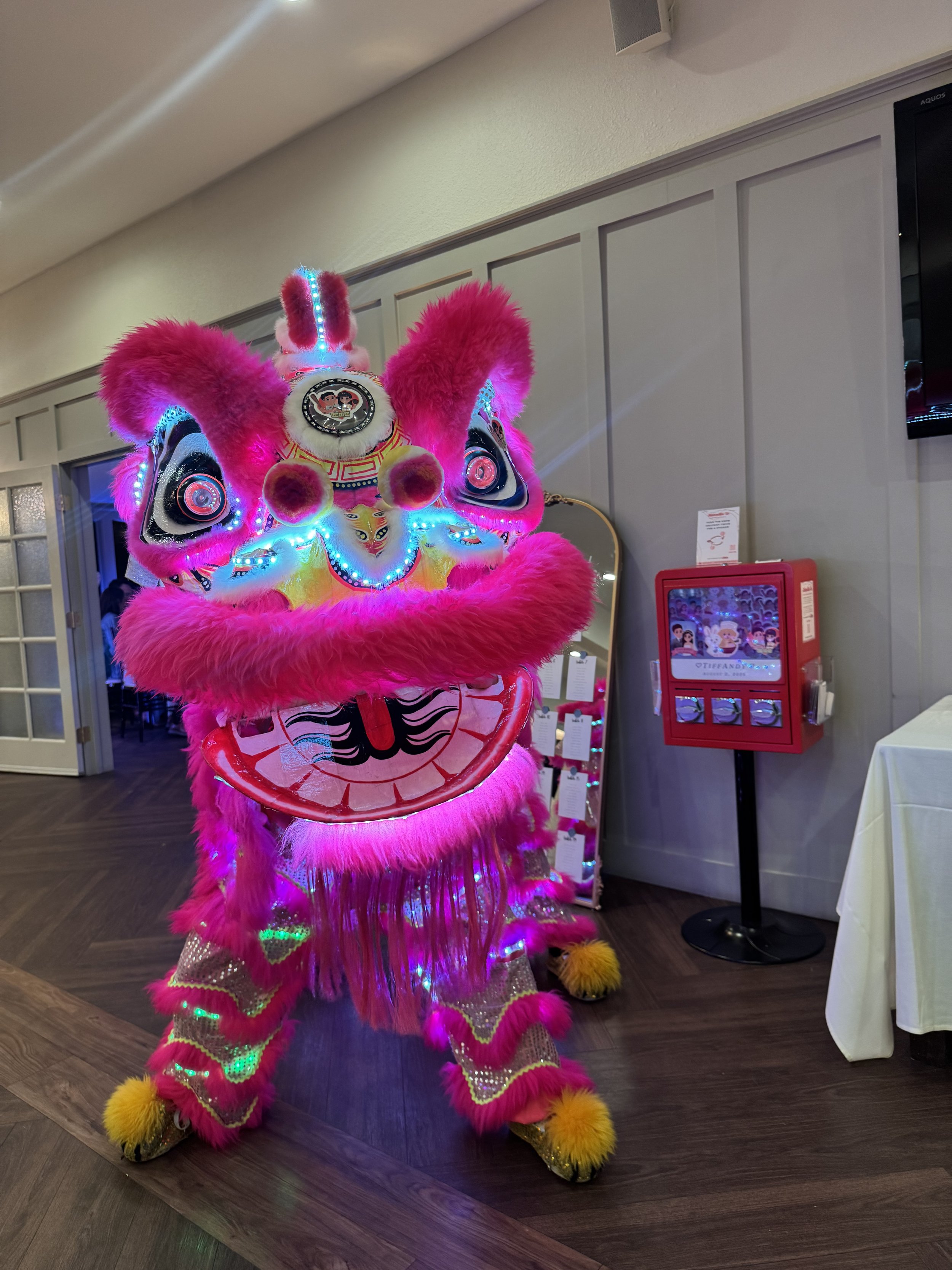 A traditional Chinese lion dance costume decorated with bright pink fur, LED lights, and intricate designs, standing indoors near a fire alarm box and a mirror.