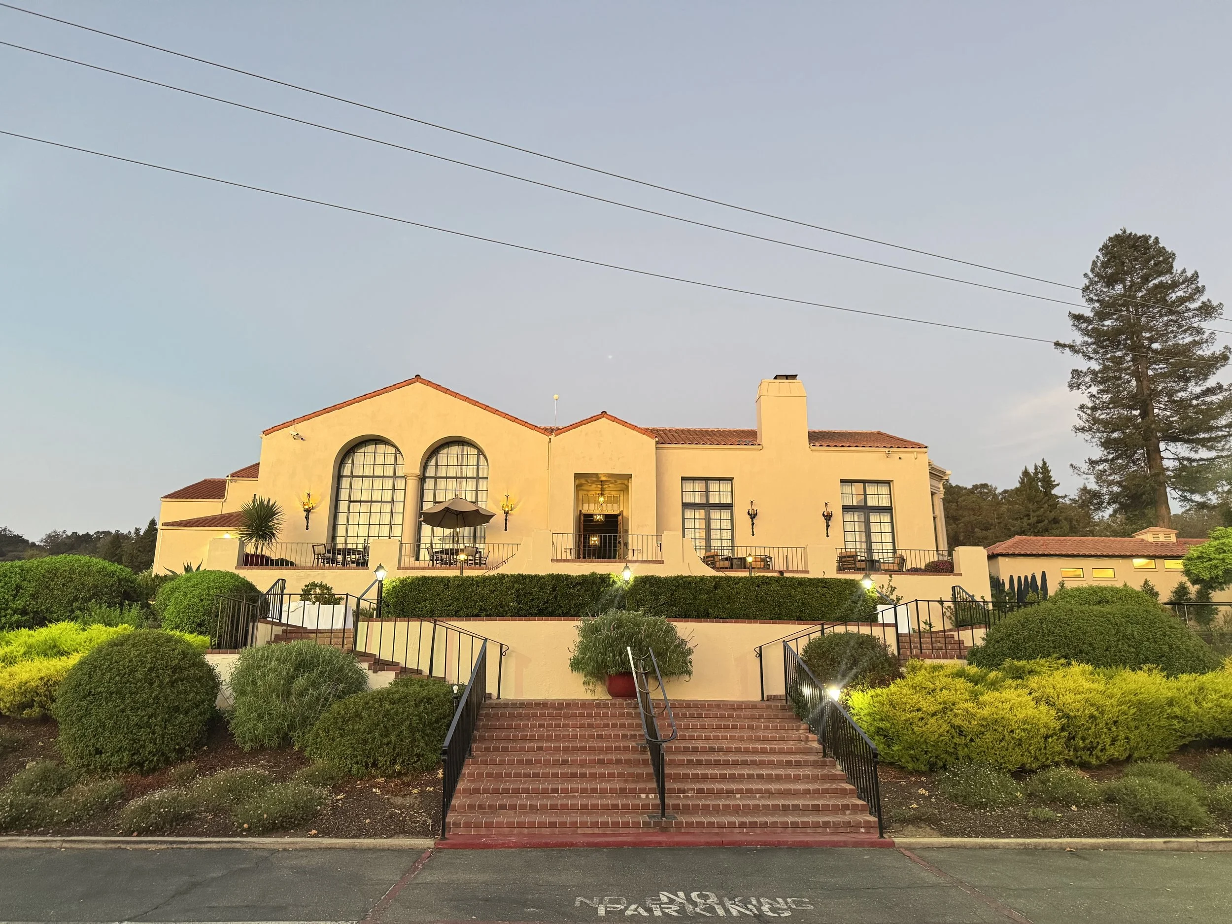 A large beige mansion with a red-tiled roof, large windows, and a front steps leading to a terrace with outdoor seating, surrounded by landscaped bushes and trees, at sunset.