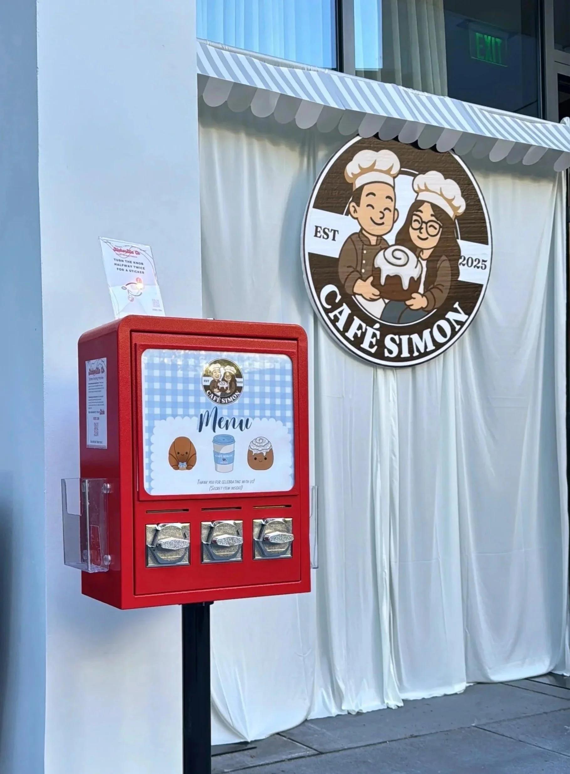 Exterior of Café Simon with a red donation or tip box in front. The box features a sign with cartoon images of a dog and a coffee cup, and a message thanking customers. The café logo, featuring cartoon characters wearing chef hats and holding a cake, is displayed on a circular sign and above the curtain.