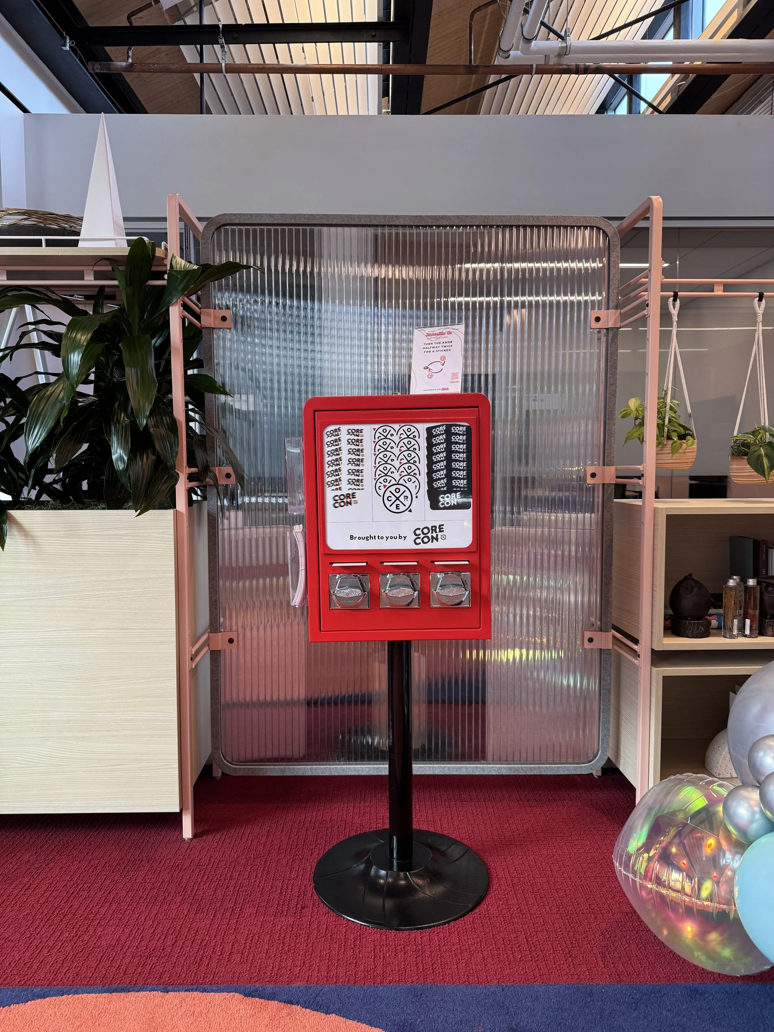 A red lottery or raffle machine with three silver levers, placed on a black stand, in front of a metal and glass partition. There are potted plants and balloons nearby, inside a modern indoor space.