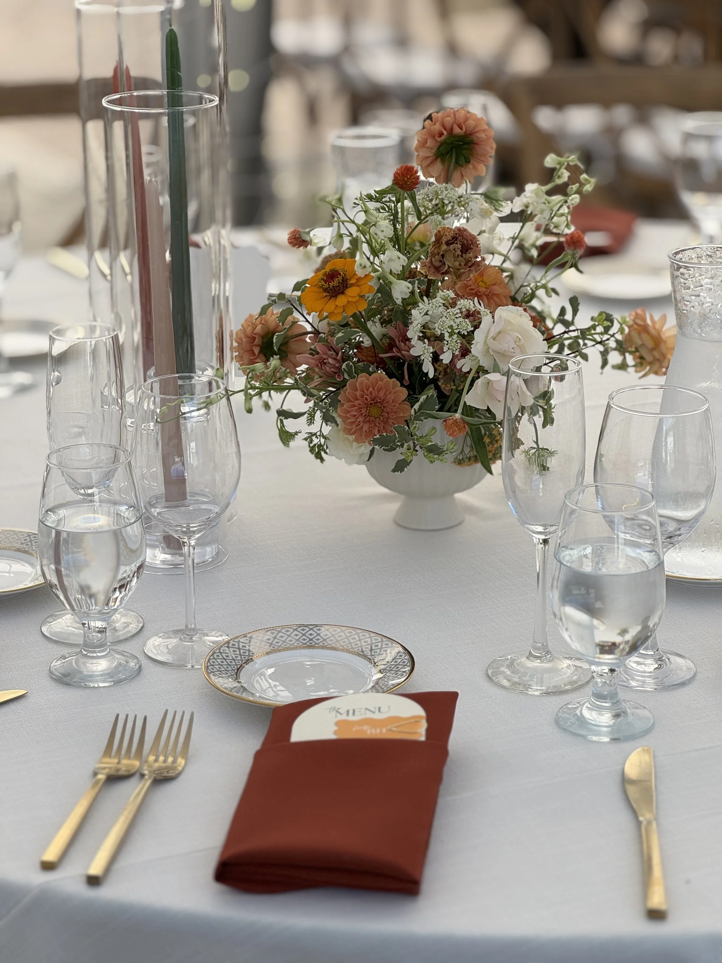 Elegant table setting with a white tablecloth, gold flatware, a folded red napkin, a menu card, glassware, and a floral centerpiece with peach, white, and orange flowers.