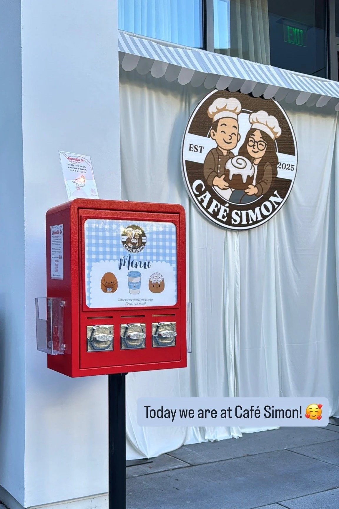 Exterior of Café Simon with a red menu box and a large round sign featuring two cartoon characters wearing chefs hats and holding a cake, set against a white backdrop. The sign and menu box display the café's logo and branding.