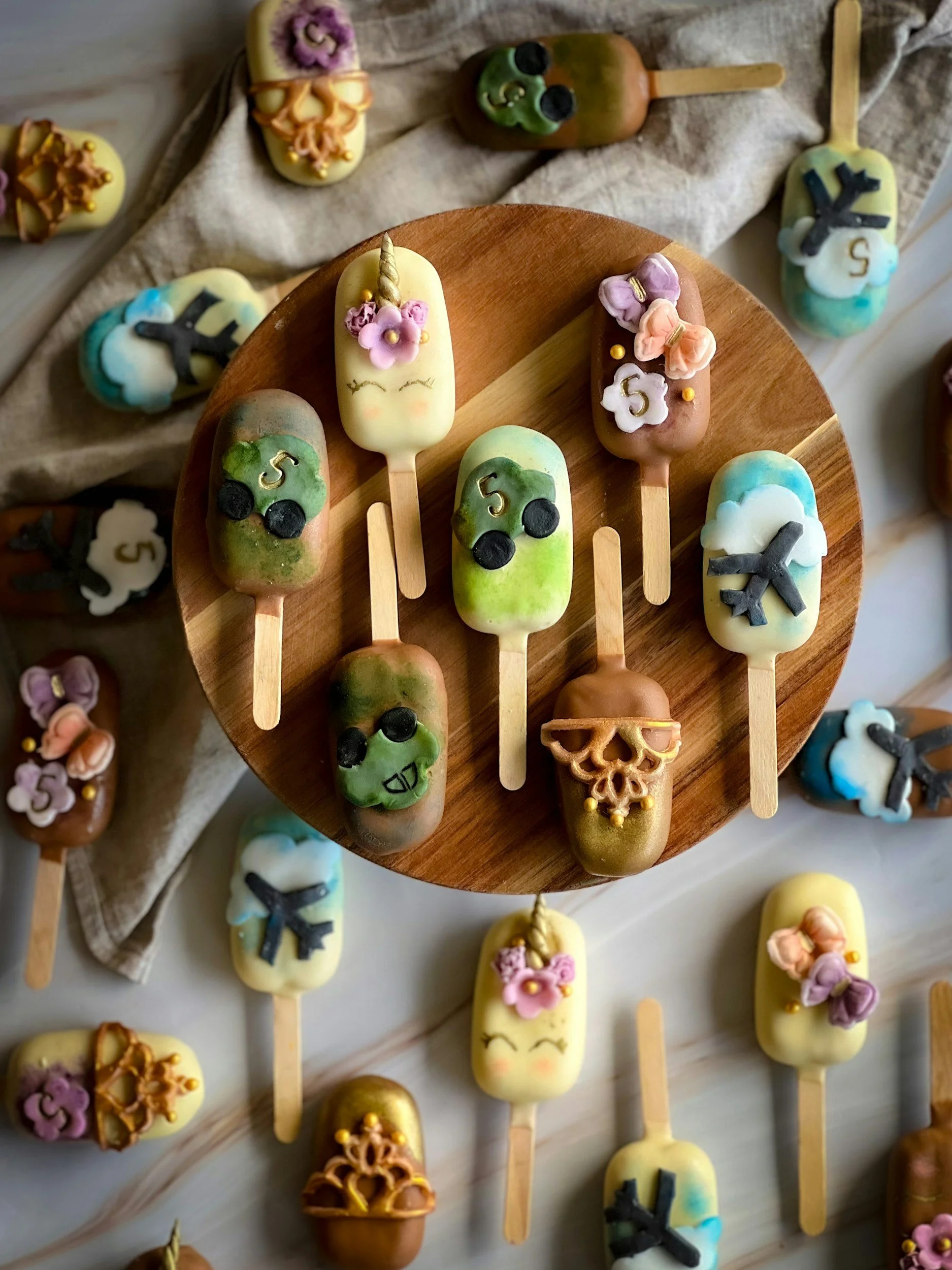 Assorted decorated popsicles arranged on a round wooden tray and scattered around, featuring various colors and Halloween-themed decorations like black cat shapes, flowers, unicorn horns, and smiley faces.