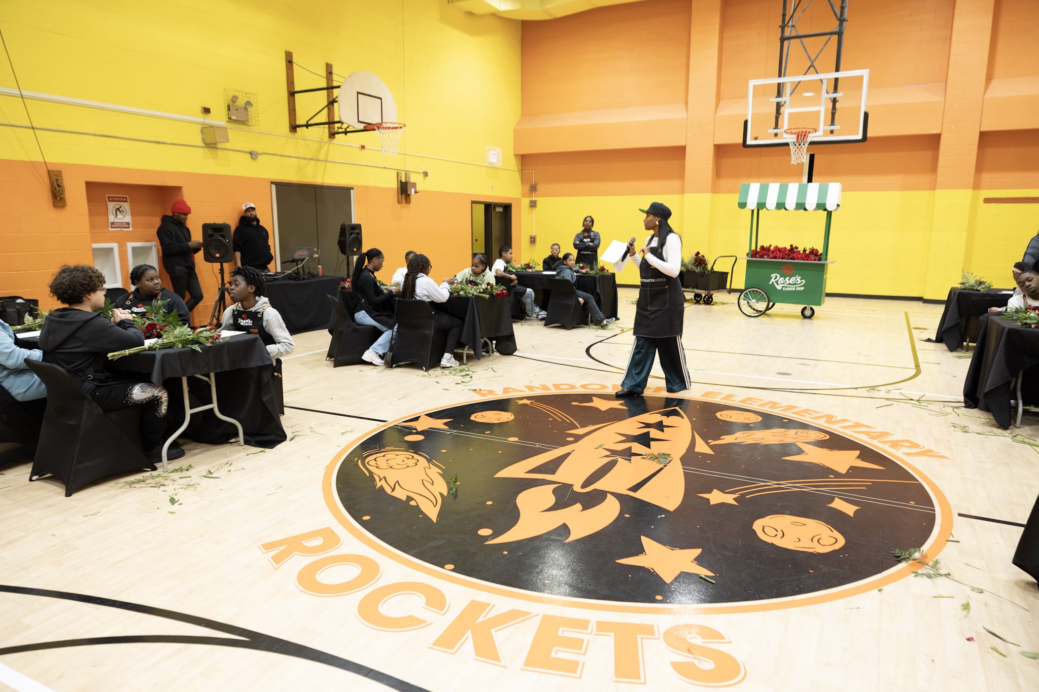 A woman stands in the center of a basketball court, speaking into a microphone during a gathering with children seated at tables covered with black tablecloths. The court has a large logo of a rocket, stars, and planets, and the walls are painted in 