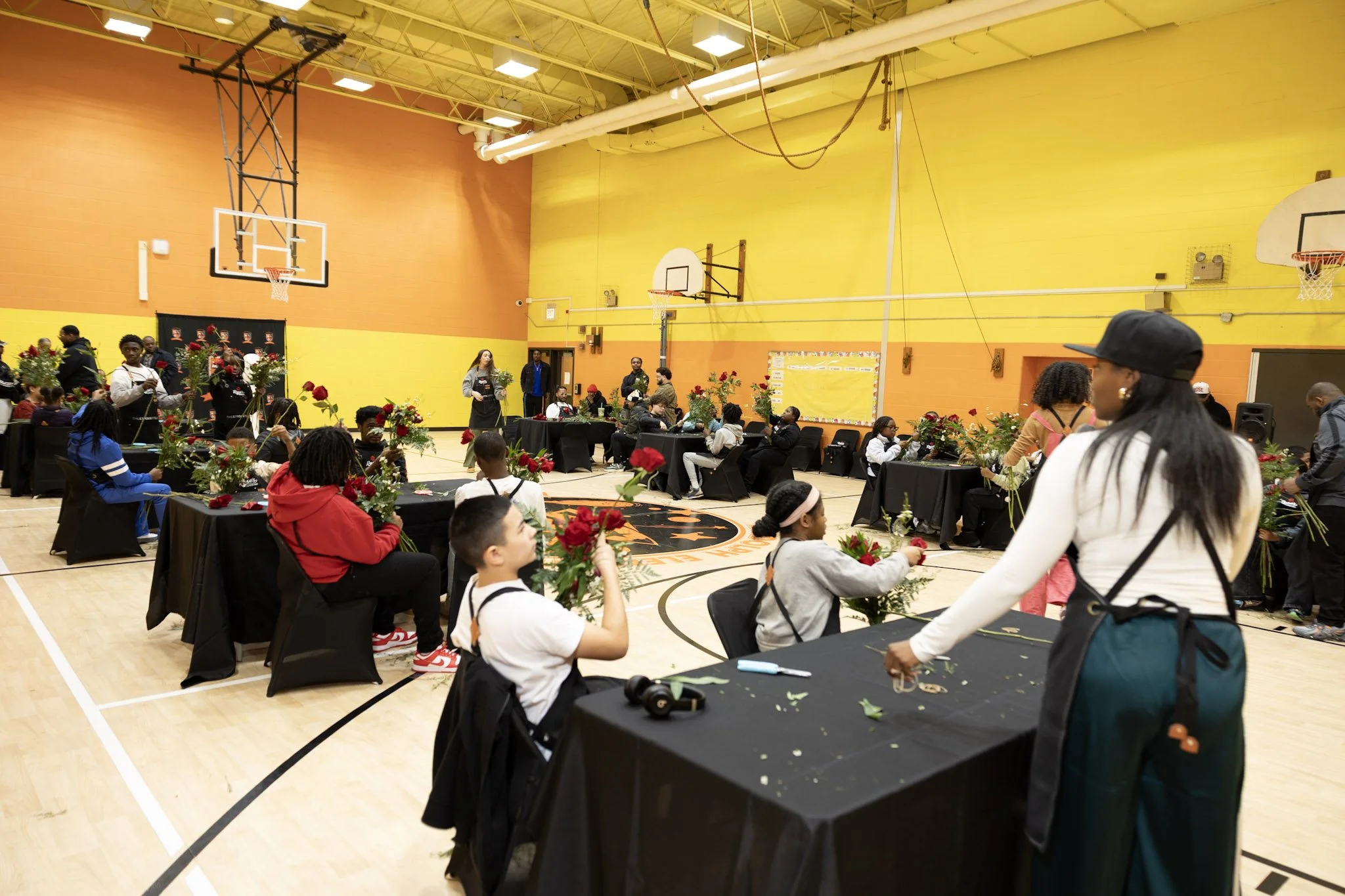 Children and adults participating in a flower arrangement class inside a gymnasium with basketball hoops, black tables, and chairs, decorated for a floral event.