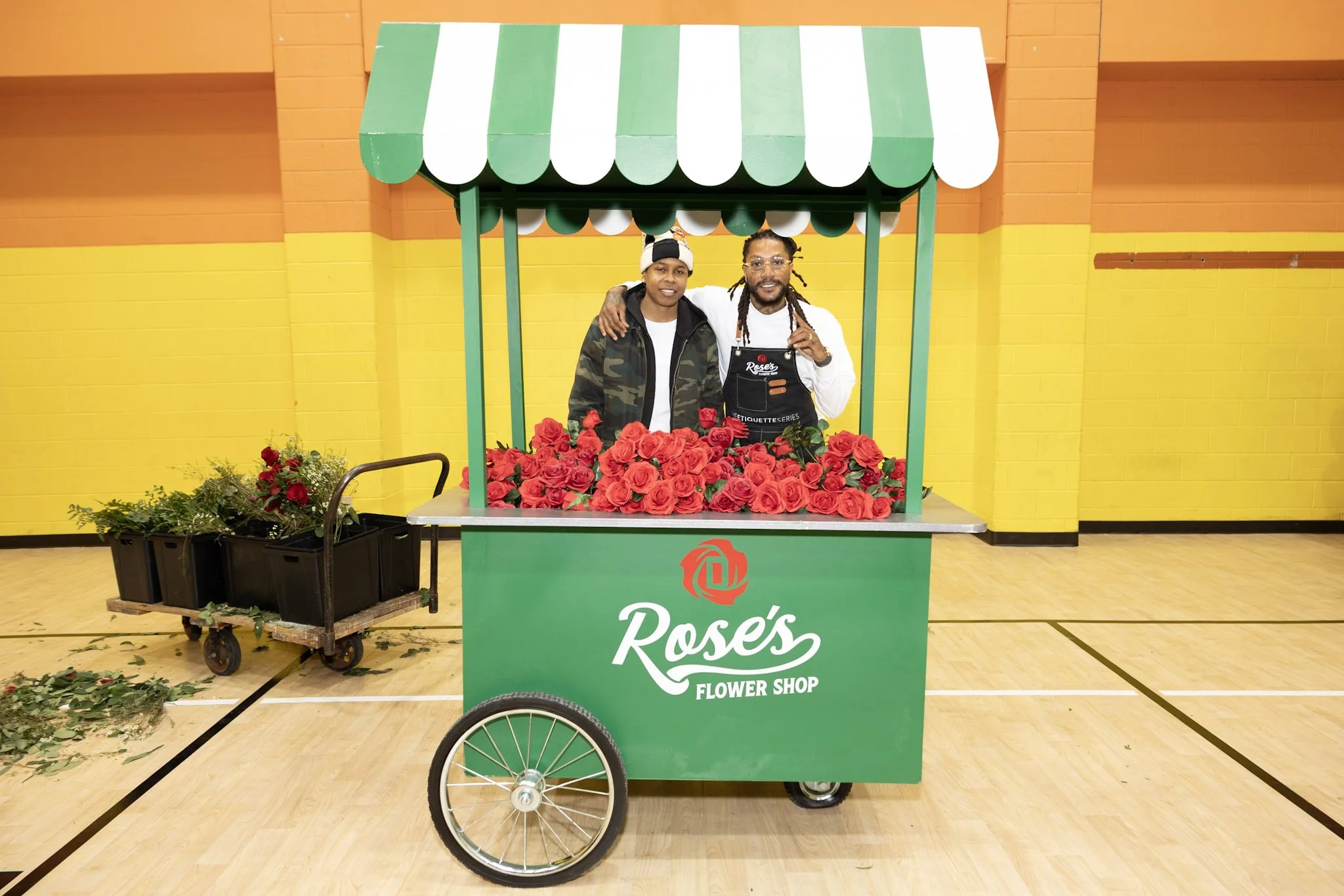 Two men standing inside a flower cart filled with red roses at an indoor event. The cart has a green and white striped canopy and a green front panel with the logo and name 'Roses Flower Shop.