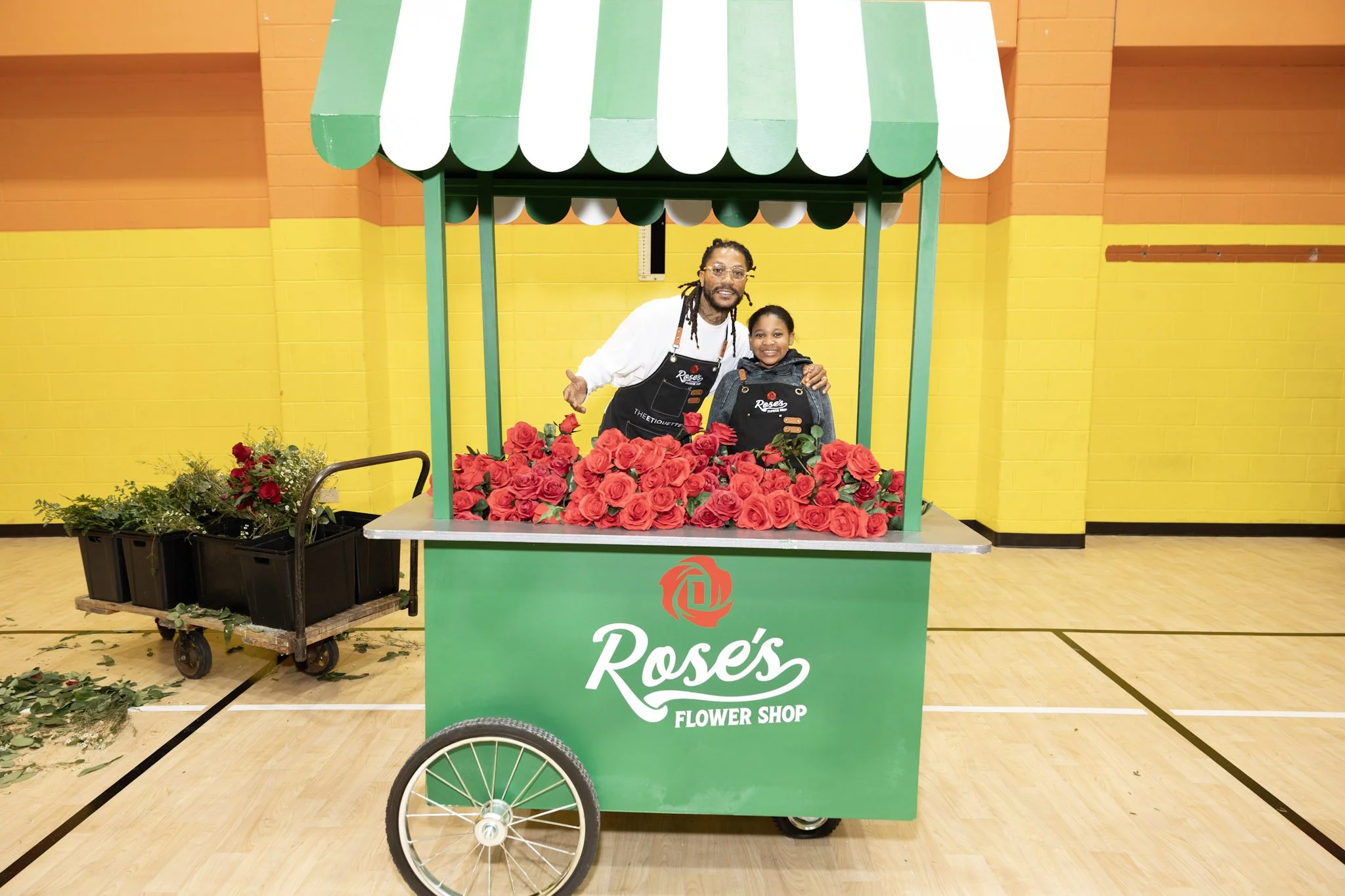 A man and a young girl standing behind a green flower cart filled with red roses, inside a gymnasium with yellow and orange walls.