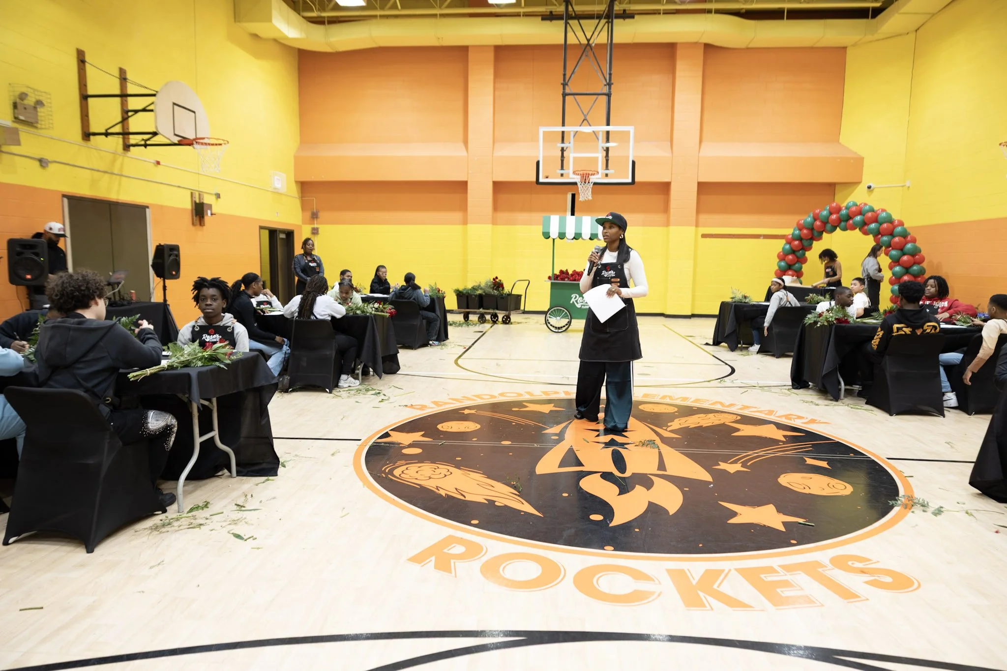 A woman standing on a basketball court in a gymnasium, speaking into a microphone during a community event. There are tables with people seated around her, some with flowers and decorations, and a balloon arch in the background. The gym has colorful 