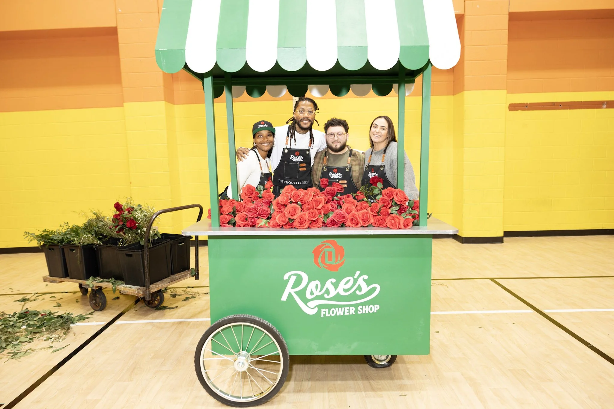 Four people inside a green flower cart with a green and white striped canopy, displaying red roses for sale, labeled 'Roses Flower Shop'. They are smiling and posing for the photo in an indoor space with bright yellow walls.