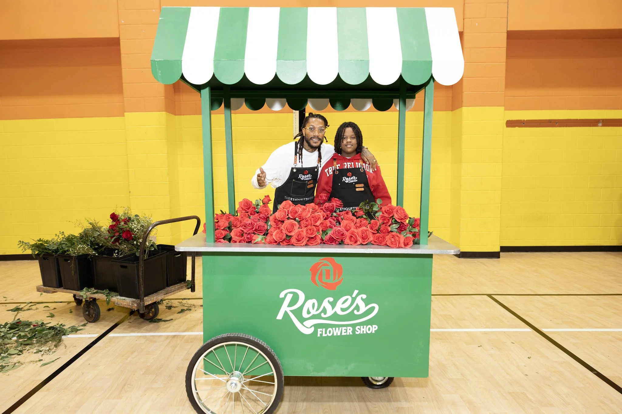 Two men standing inside a flower cart filled with pink roses at Rose's Flower Shop. The cart has a green and white striped canopy and is situated indoors with a bright yellow and orange brick wall in the background.