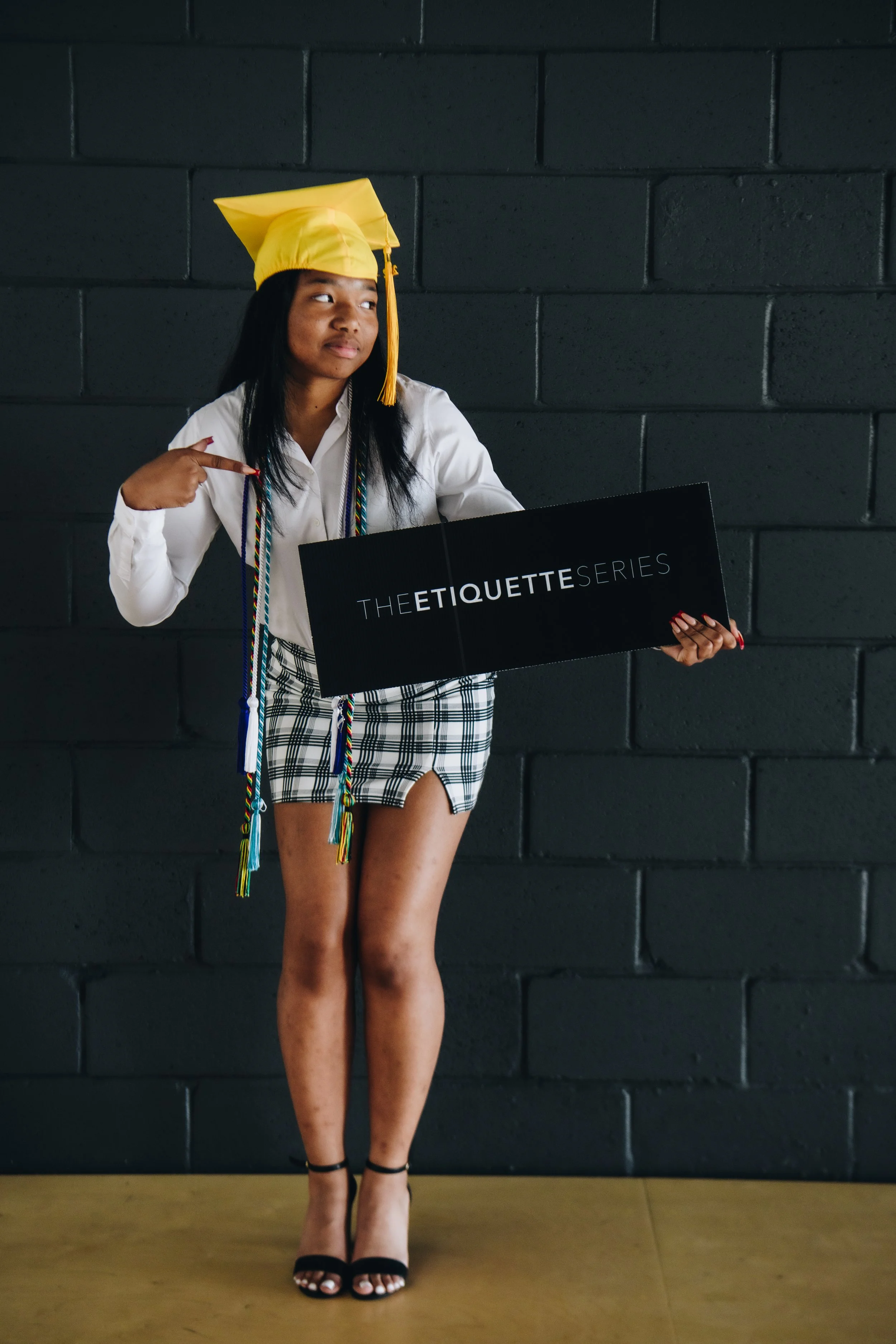 Young woman in a yellow graduation cap and plaid skirt holding a black sign that reads "The Etiquette Series" against a black brick wall. The Etiquette Series, Chicago-Detroit.