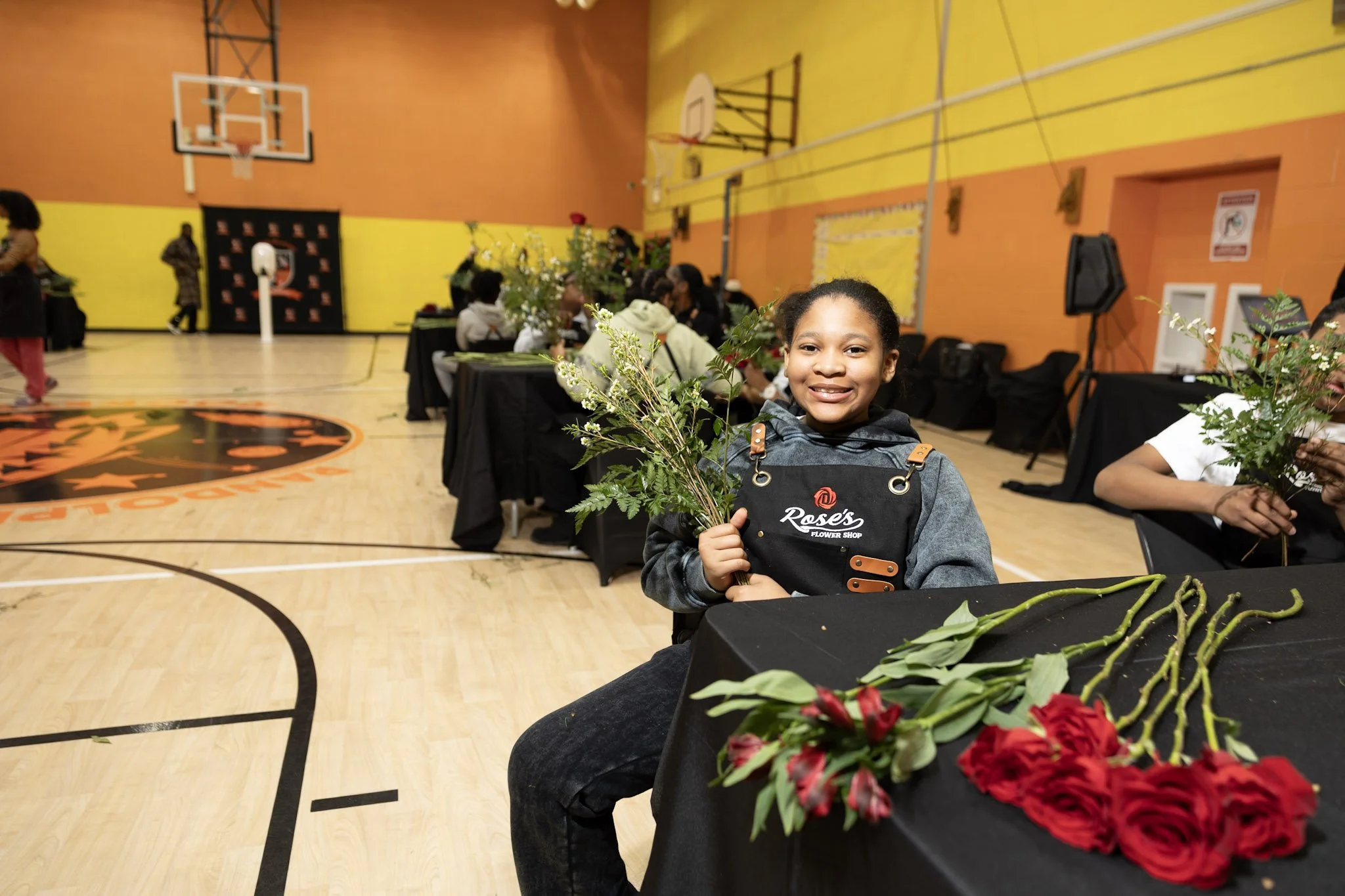 A young girl smiling and holding a bouquet of white flowers in a gymnasium with tables of flowers, and other children participating in a flower arrangement event.