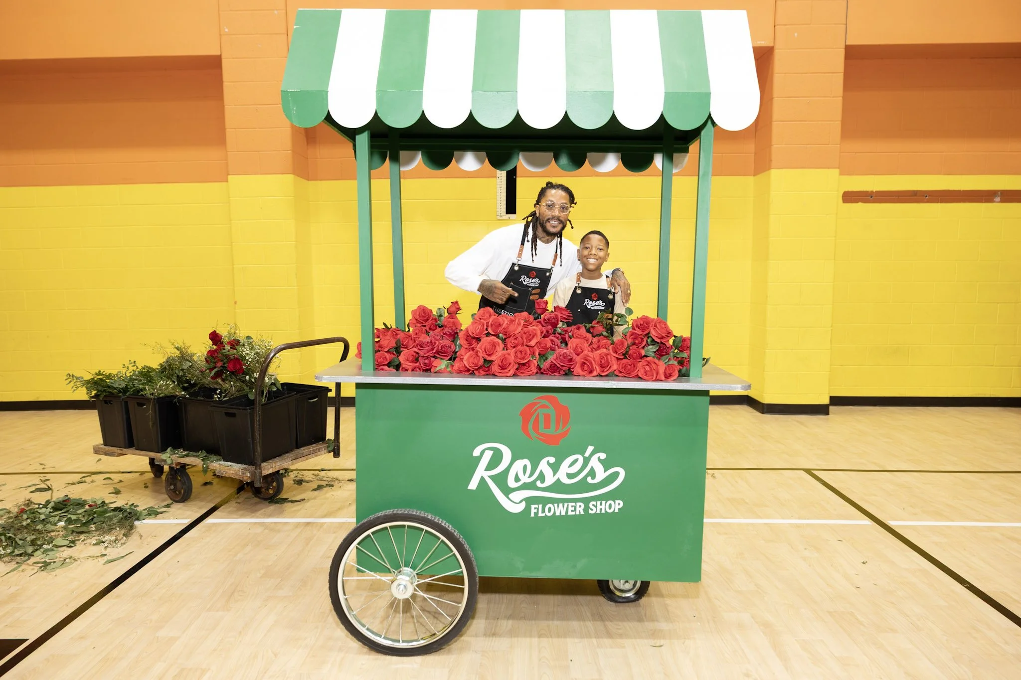 Two people standing behind a green flower cart labeled "Rose's Flower Shop" with red roses on top, inside a gymnasium with yellow and orange walls.