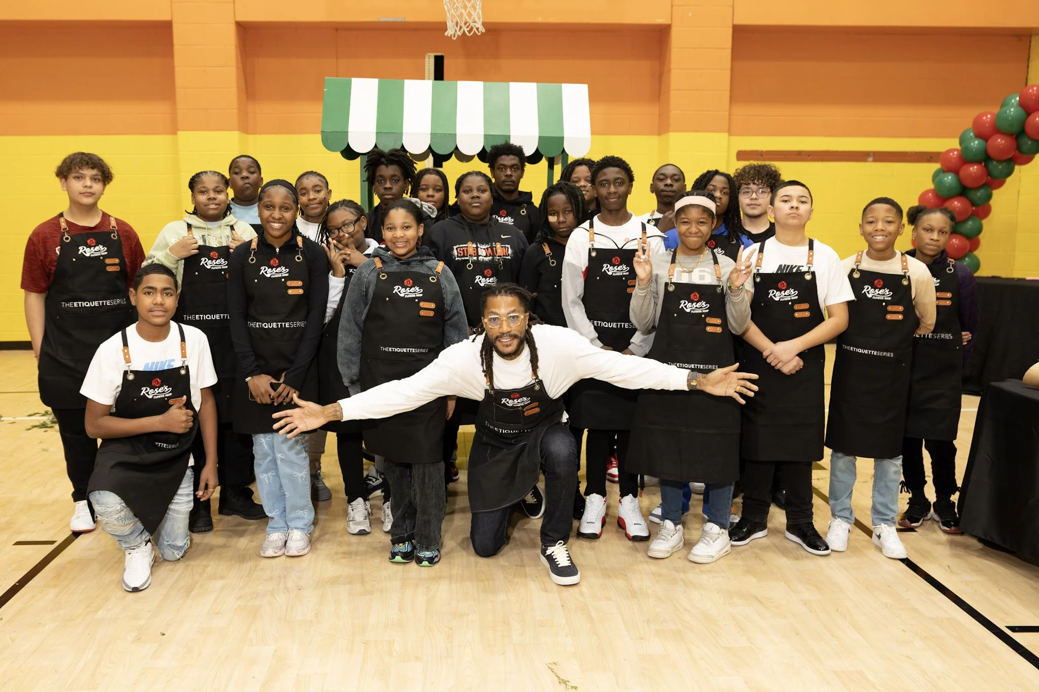 A group of young students and their instructor posing in a gymnasium. The students are wearing black aprons with a logo, and the instructor is kneeling in the front with arms wide open. The background features bright yellow and orange walls, a green 