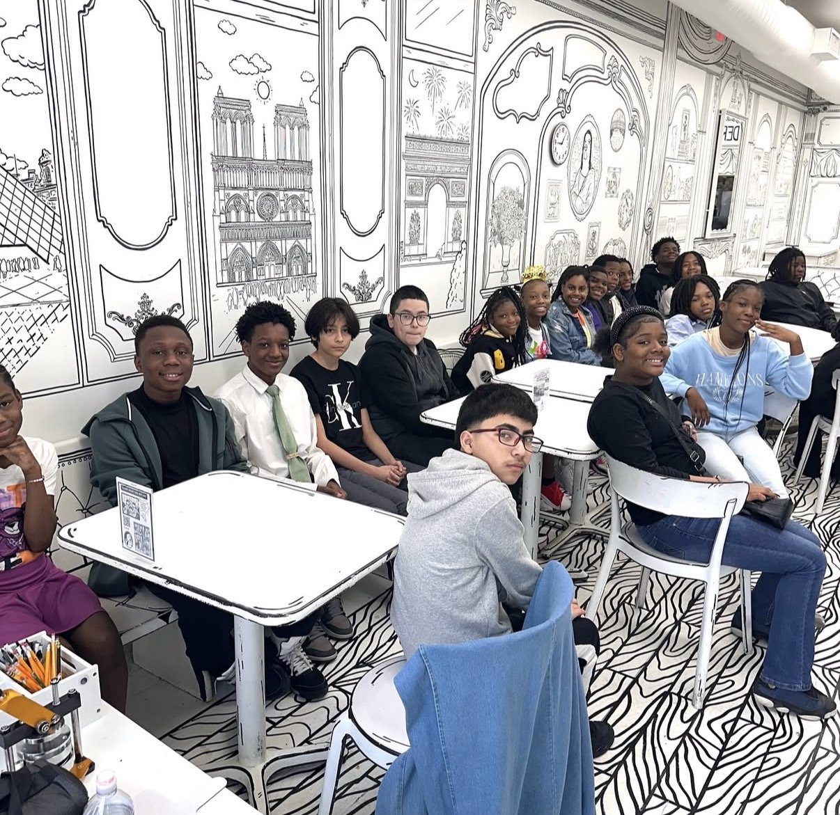 A group of diverse children and teenagers sitting at tables in a classroom with black and white mural wall art depicting Paris landmarks. The Etiquette Series, Chicago-Detroit.