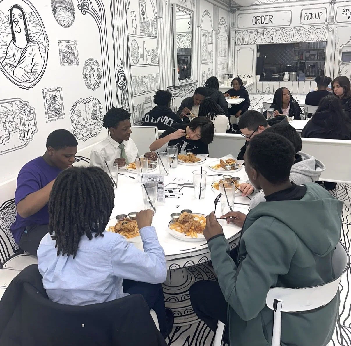 Group of young people dining together at a restaurant with black-and-white wall art resembling line drawings of classic art and decor. The Etiquette Series, Chicago-Detroit.
