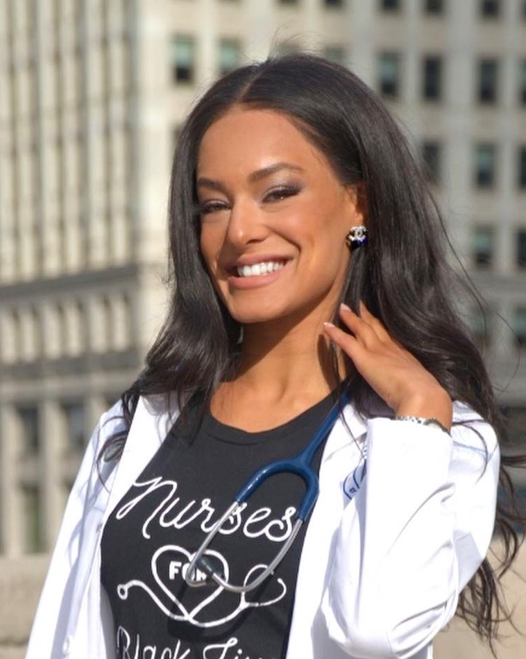 A woman with long black hair smiling outdoors, wearing a white coat, a black t-shirt with white text, and a stethoscope around her neck, with city buildings in the background.