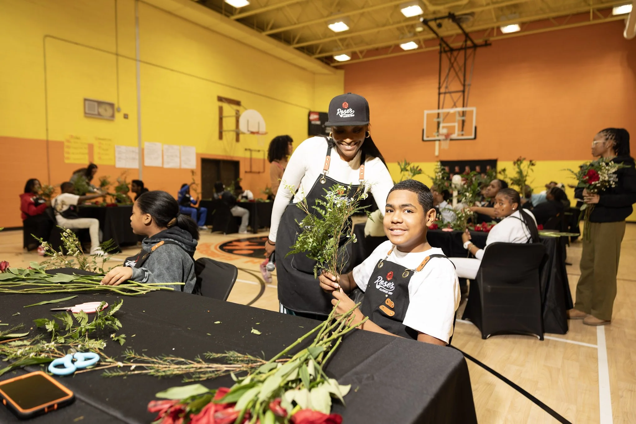 A young boy sitting at a table holding flowers, smiling, with a woman leaning over him placing her hand on his shoulder. The group appears to be in a gymnasium with other children and adults, some holding flowers, participating in a flower arranging 