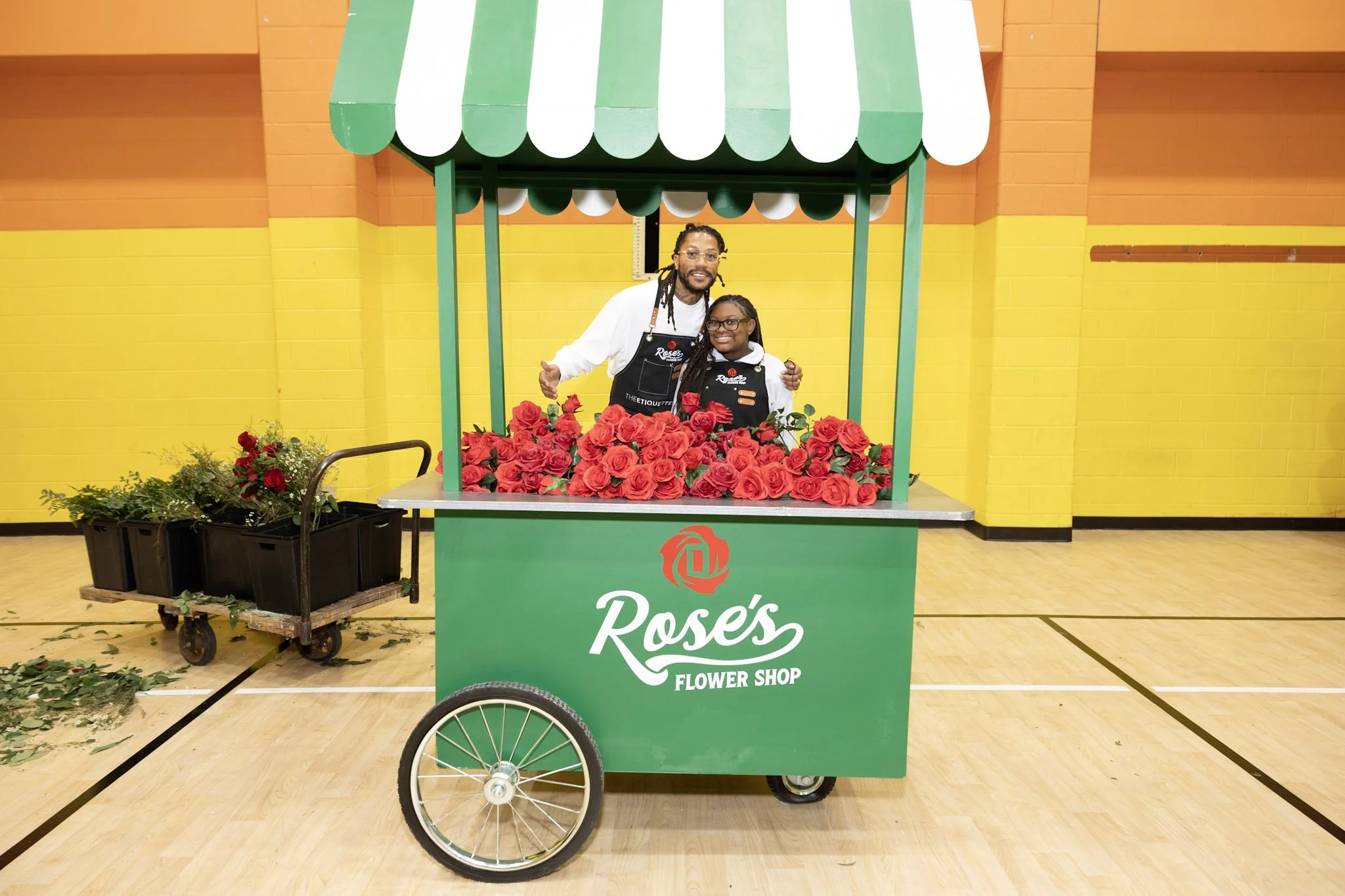 Two people standing behind a cart filled with red roses at Rose's Flower Shop, inside a gymnasium with yellow and orange walls.