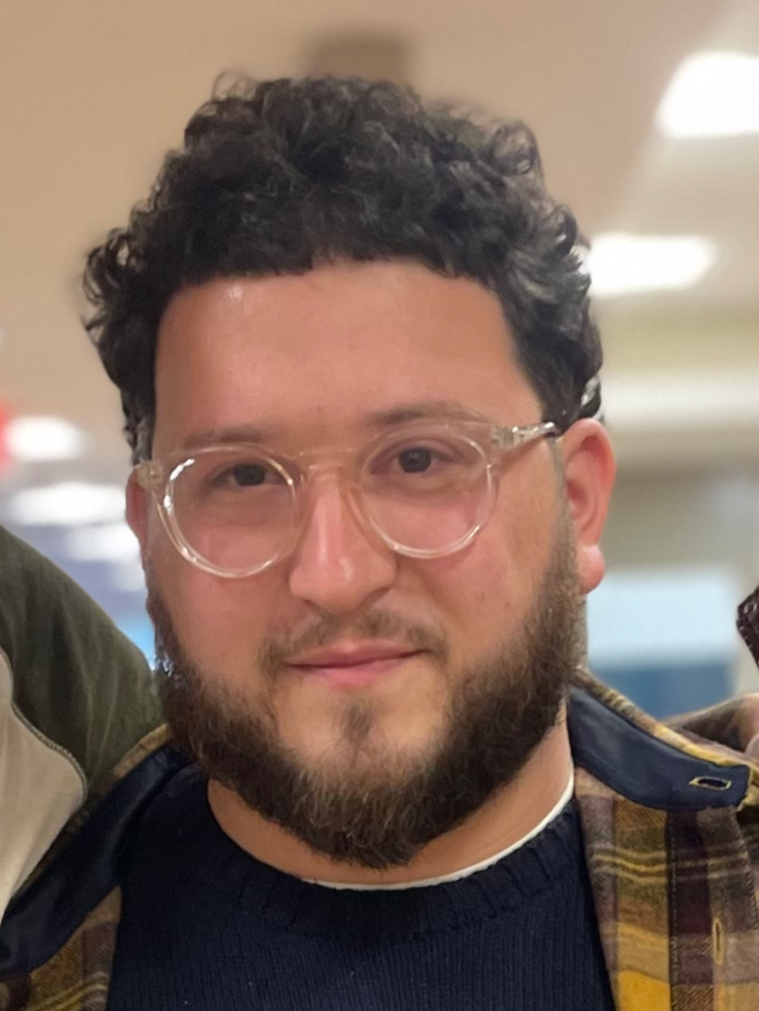 A man with curly dark hair, clear glasses, a beard, and wearing a dark sweater and plaid shirt, smiling indoors.