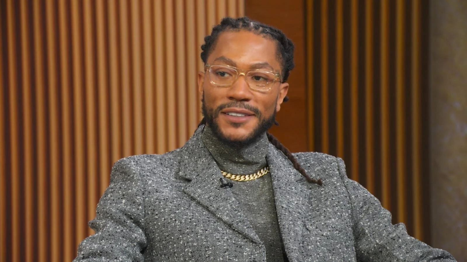 A man with glasses and dreadlocks wearing a sparkly gray blazer and a chain necklace, smiling in a room with wooden paneling background.