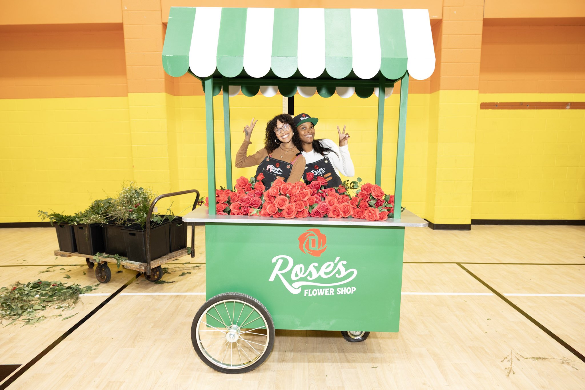 Two women in flower shop uniforms smiling and making peace signs behind a green flower cart filled with pink roses, with a yellow and orange wall in the background.