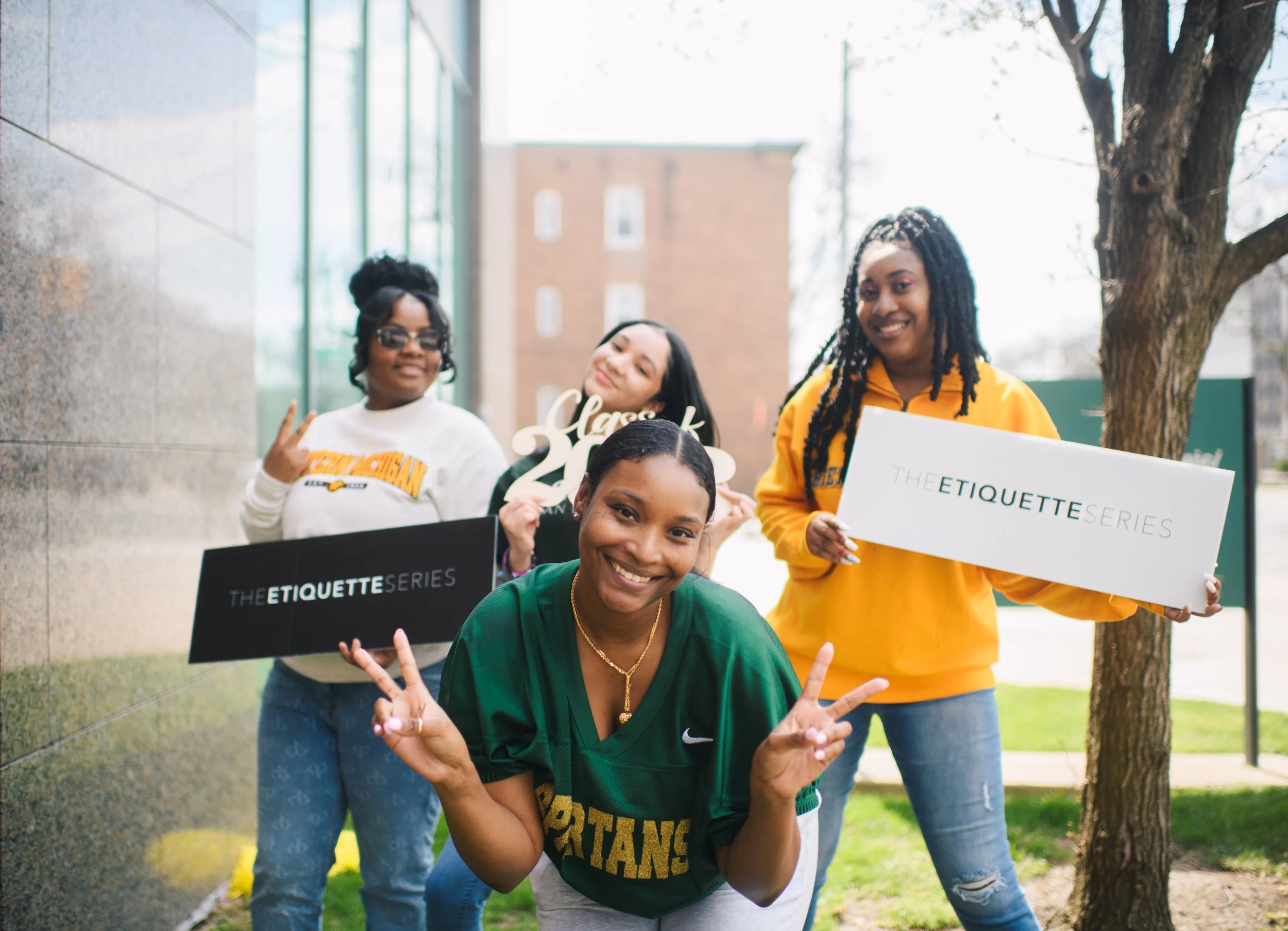Group of five women outside, smiling, making peace signs, holding signs that say "The Etiquette Series" and "Class of 2023," with a background of a tree, grass, and a building.
