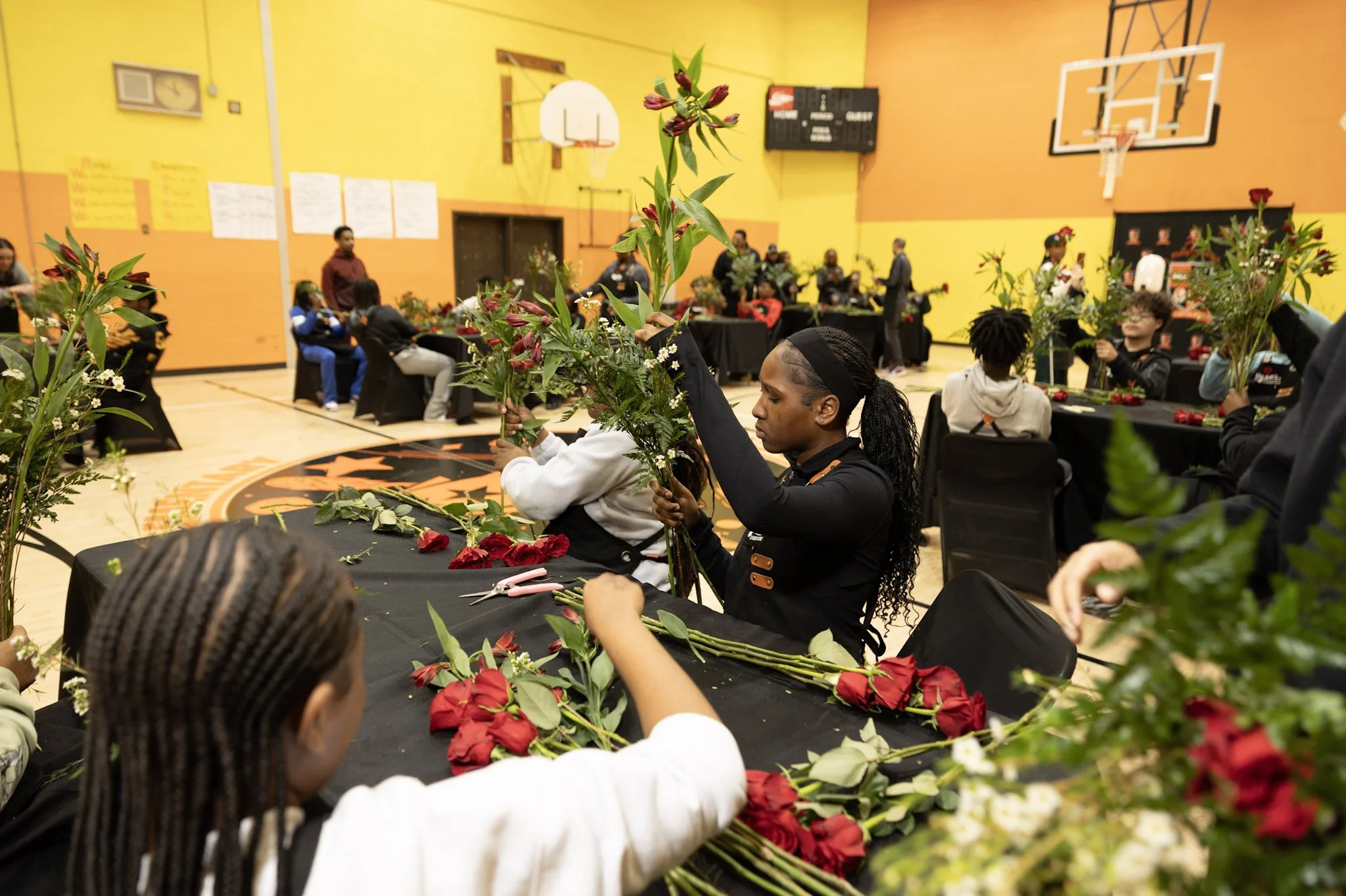 People creating floral arrangements at tables in a gymnasium with orange and yellow walls, basketball hoops, and a black logo on the floor.