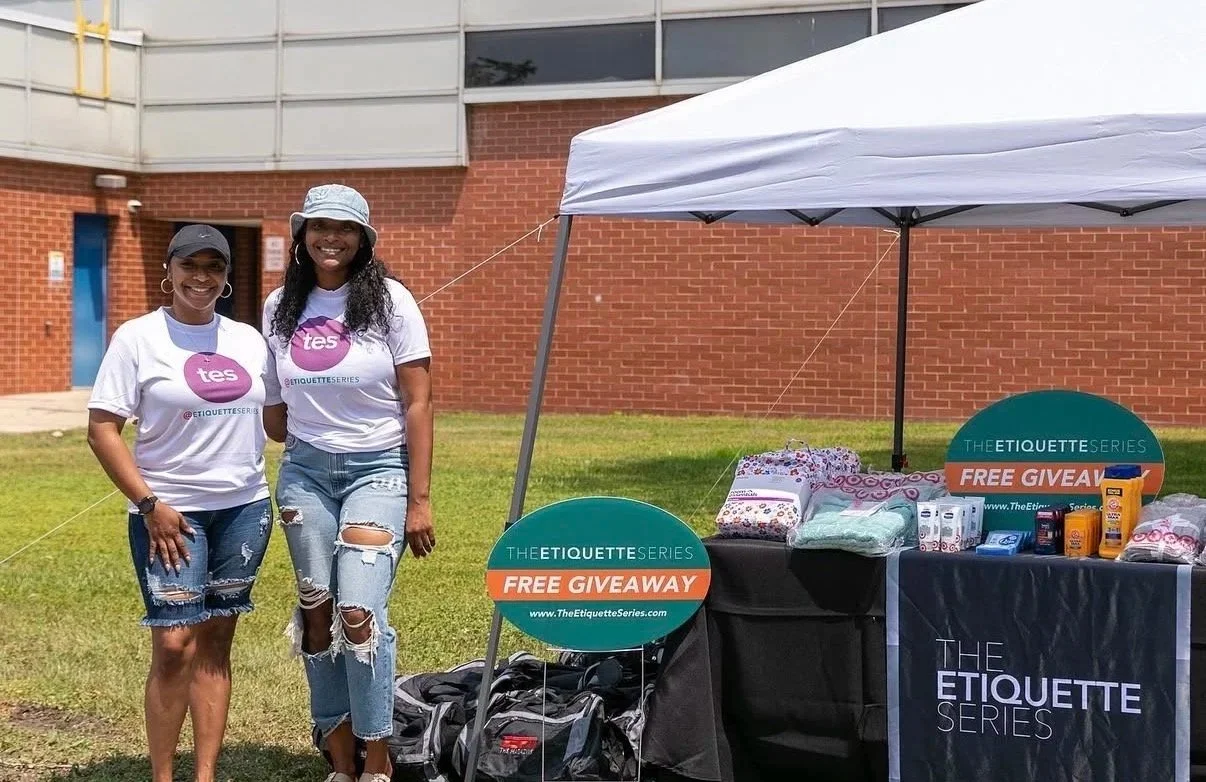 Two women standing outdoors at a promotional event booth for The Etiquette Series, with signs advertising a free giveaway and a table displaying various products.