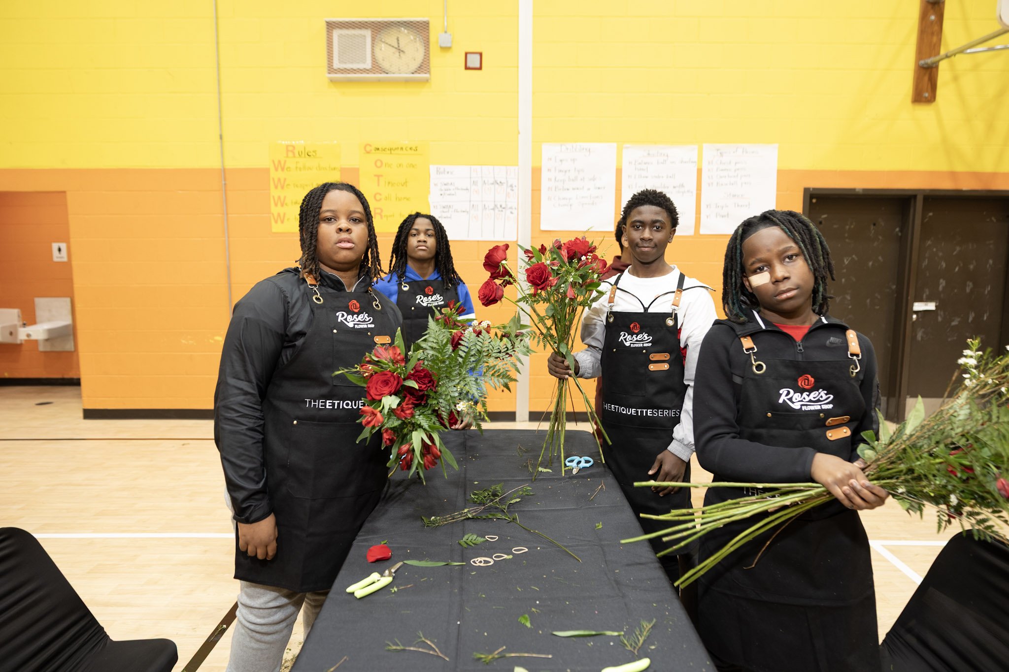 Four children wearing black aprons, standing behind a table with flower arrangements and tools, in a brightly colored indoor space with yellow and orange walls.