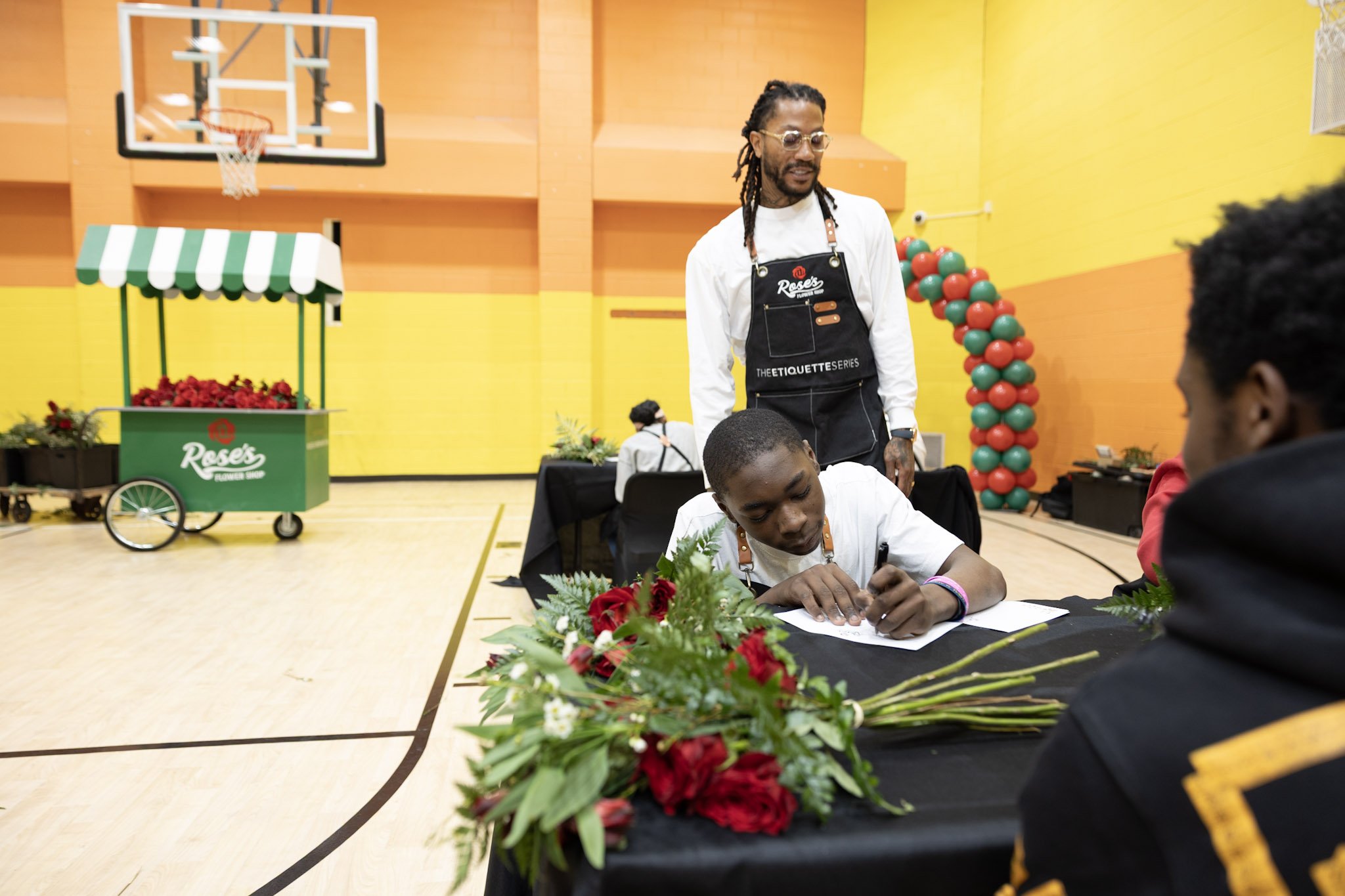 A man is signing a document on a table decorated with red roses, while another person watches. Behind him, a man with glasses and dreadlocks stands. They are in a gymnasium decorated with balloons and a strawberry stand in the background.
