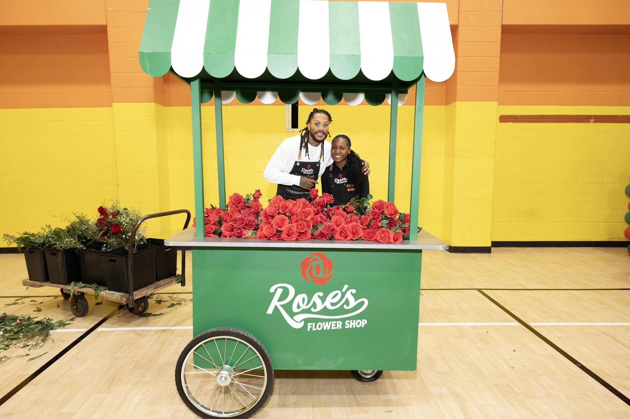 Two people standing inside a green flower cart labeled 'Rose's Flower Shop' filled with red roses, in an indoor gymnasium with yellow walls.