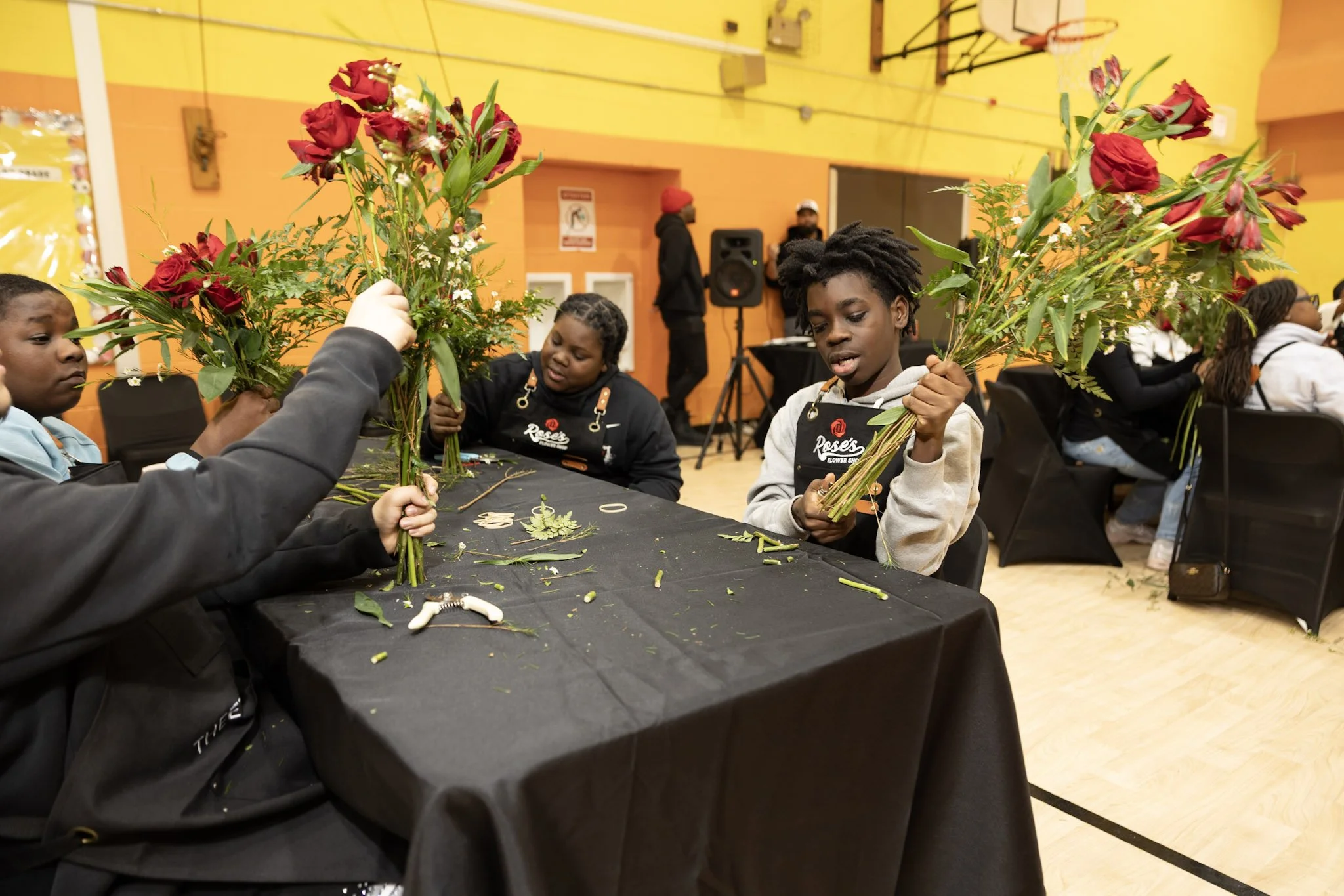 Children sitting at a table assembling large bouquets of red roses with green foliage at an indoor event.