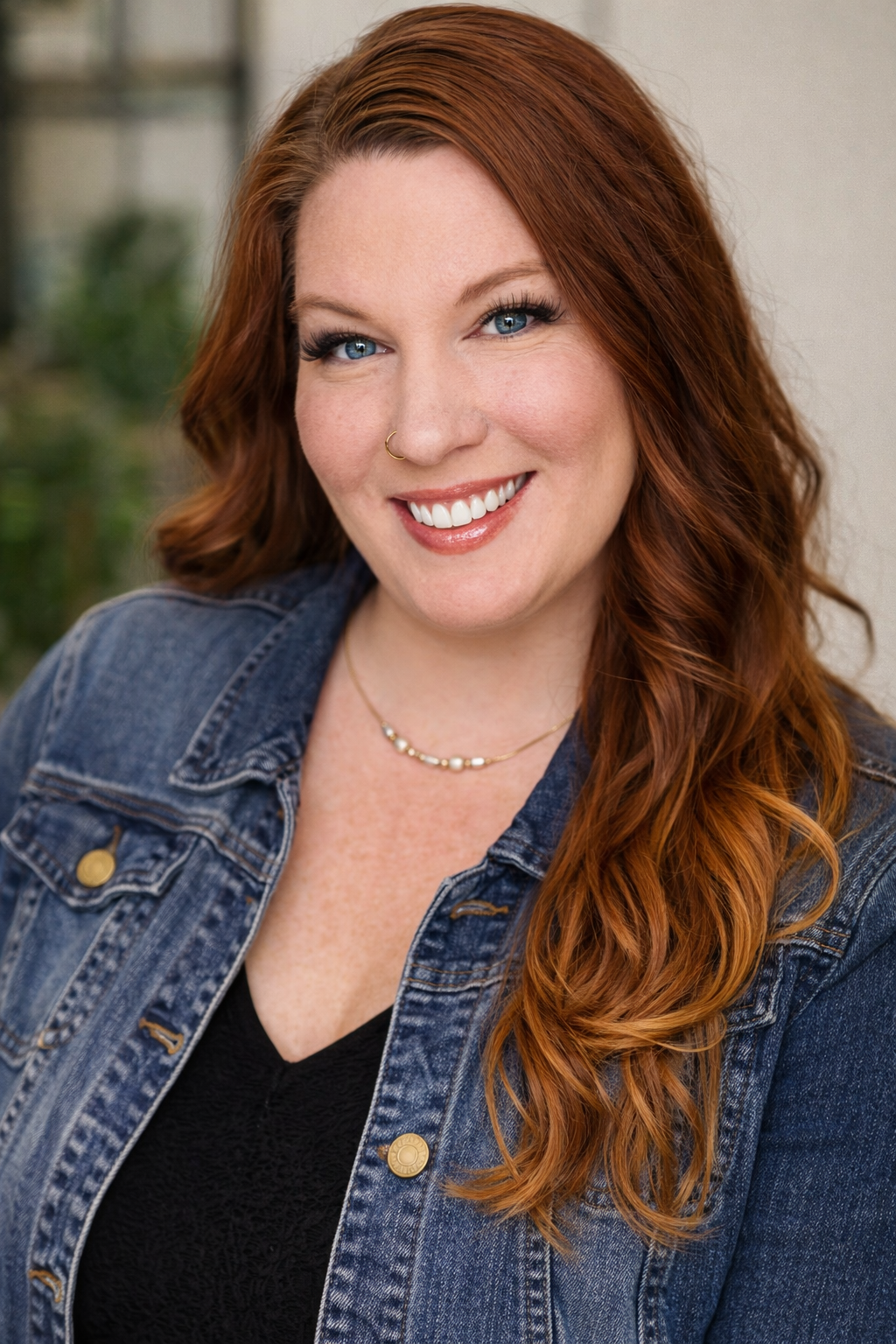 Ashley with red hair, blue eyes, and a nose ring, wearing a denim jacket and gold jewelry, in an indoor setting.